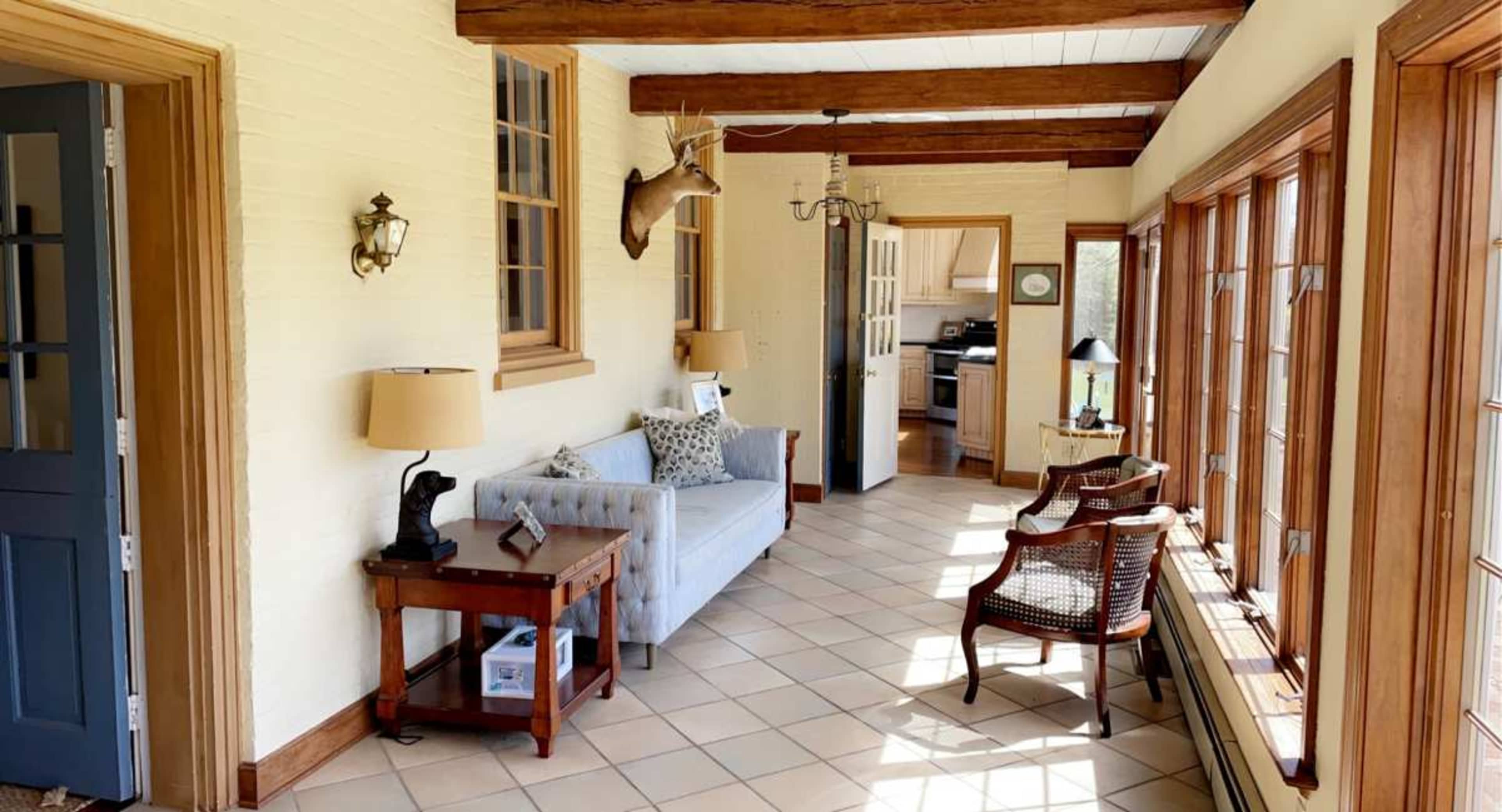 The image shows a sunlit hallway with a couch, wooden beams, a deer mount on the wall, and large windows.