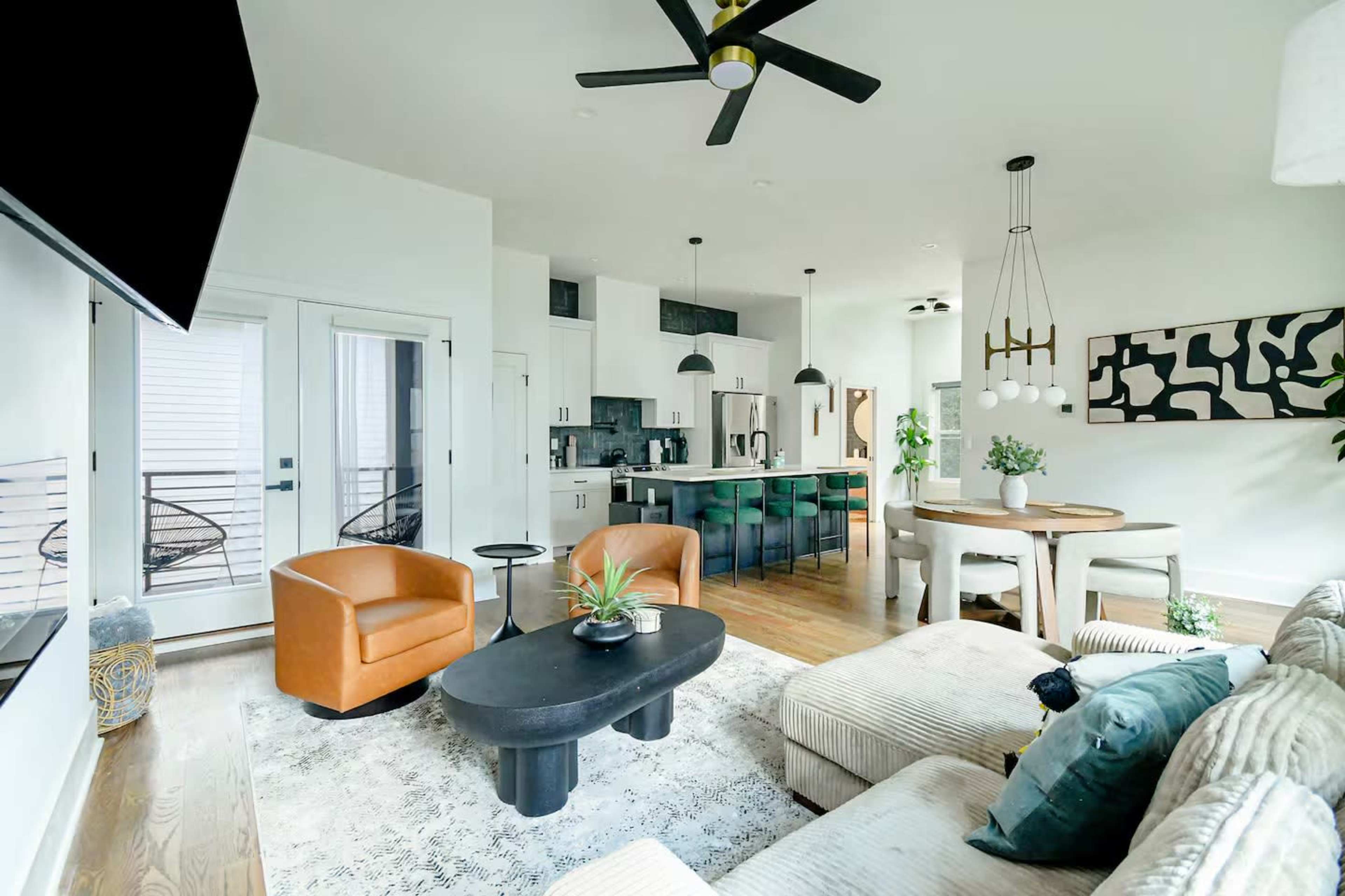The image shows a modern living room and kitchen area with a large sofa, a round dining table, and a kitchen island featuring bar stools.