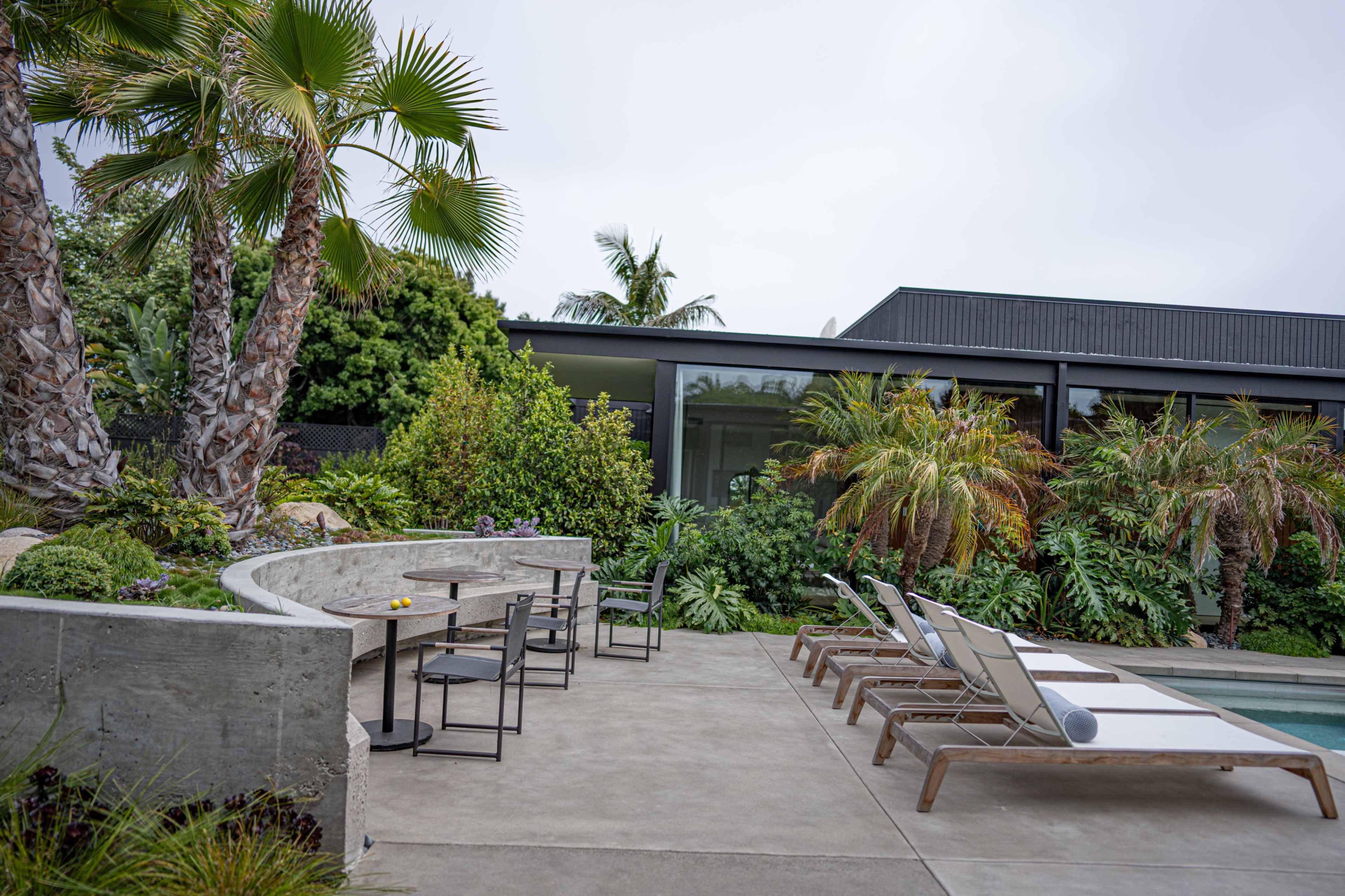 A modern outdoor area features a poolside seating arrangement with loungers and a concrete table set amidst lush tropical landscaping.