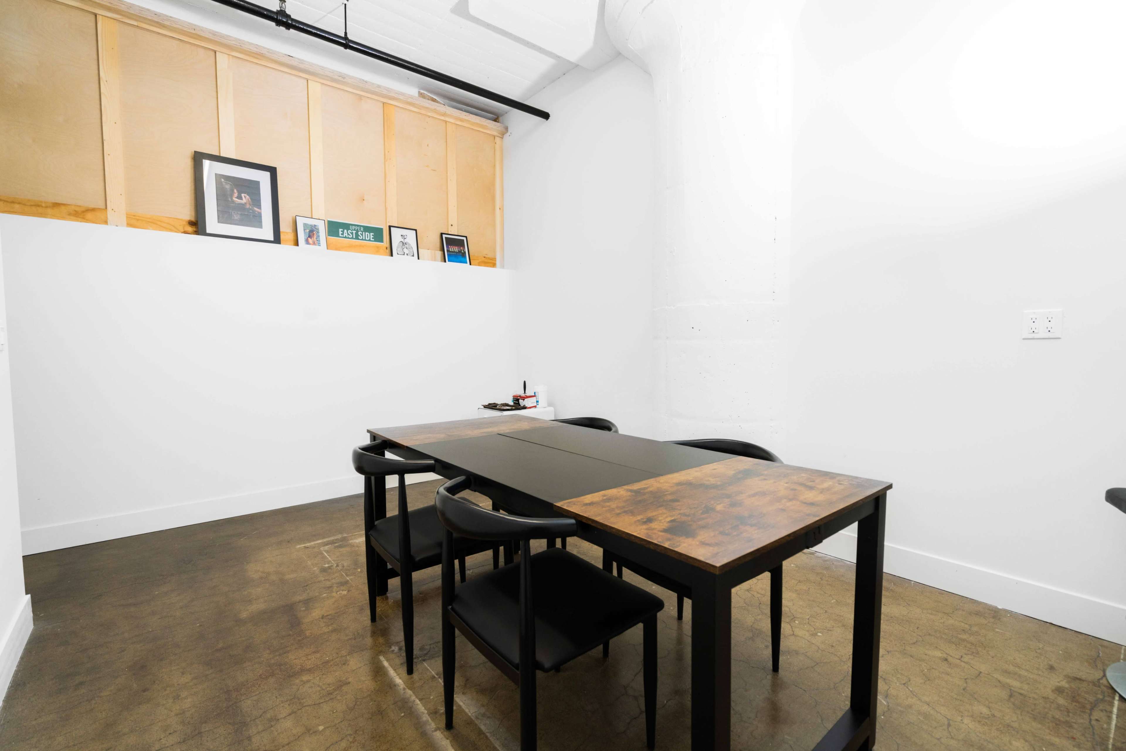 A minimalist meeting room features a long table surrounded by four black chairs against a white wall with wooden paneling and framed pictures above.