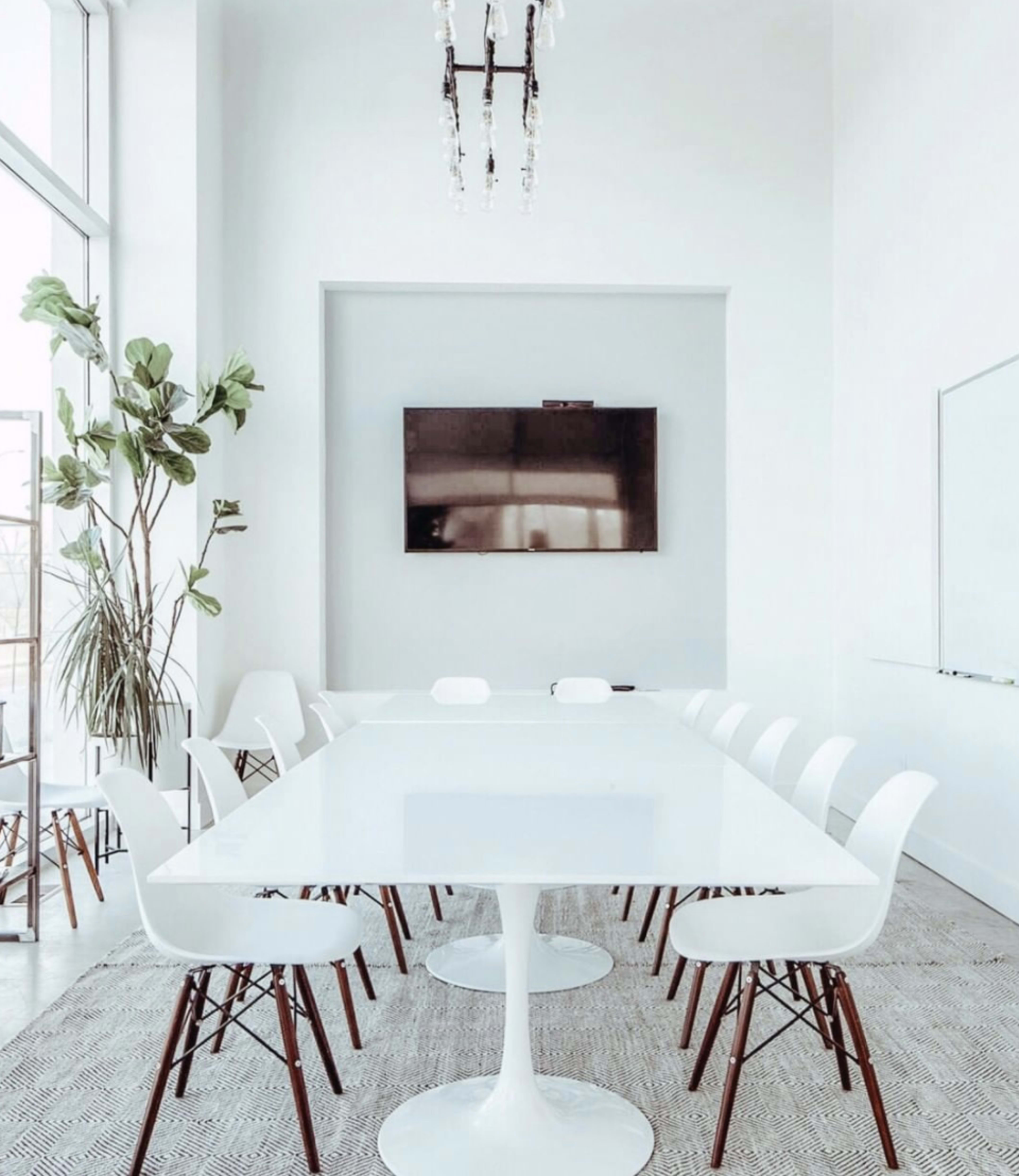 A modern conference room features a large white table surrounded by white chairs, with a television mounted on the wall and natural light streaming through large windows.