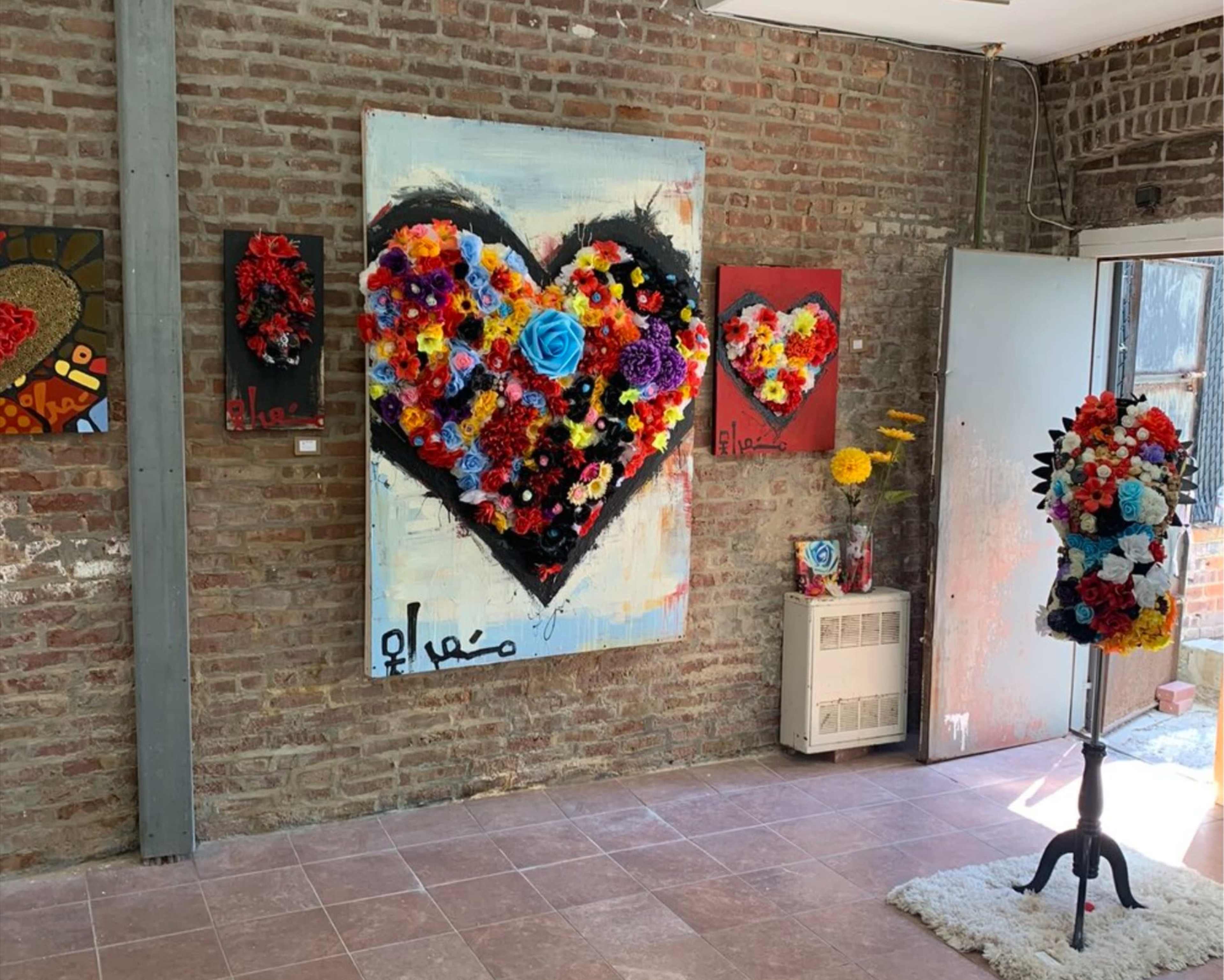 The scene displays a gallery space featuring vibrant floral artwork, including a large heart piece on the main wall and smaller pieces alongside, with a decorative sculpture adorned with flowers in the foreground.