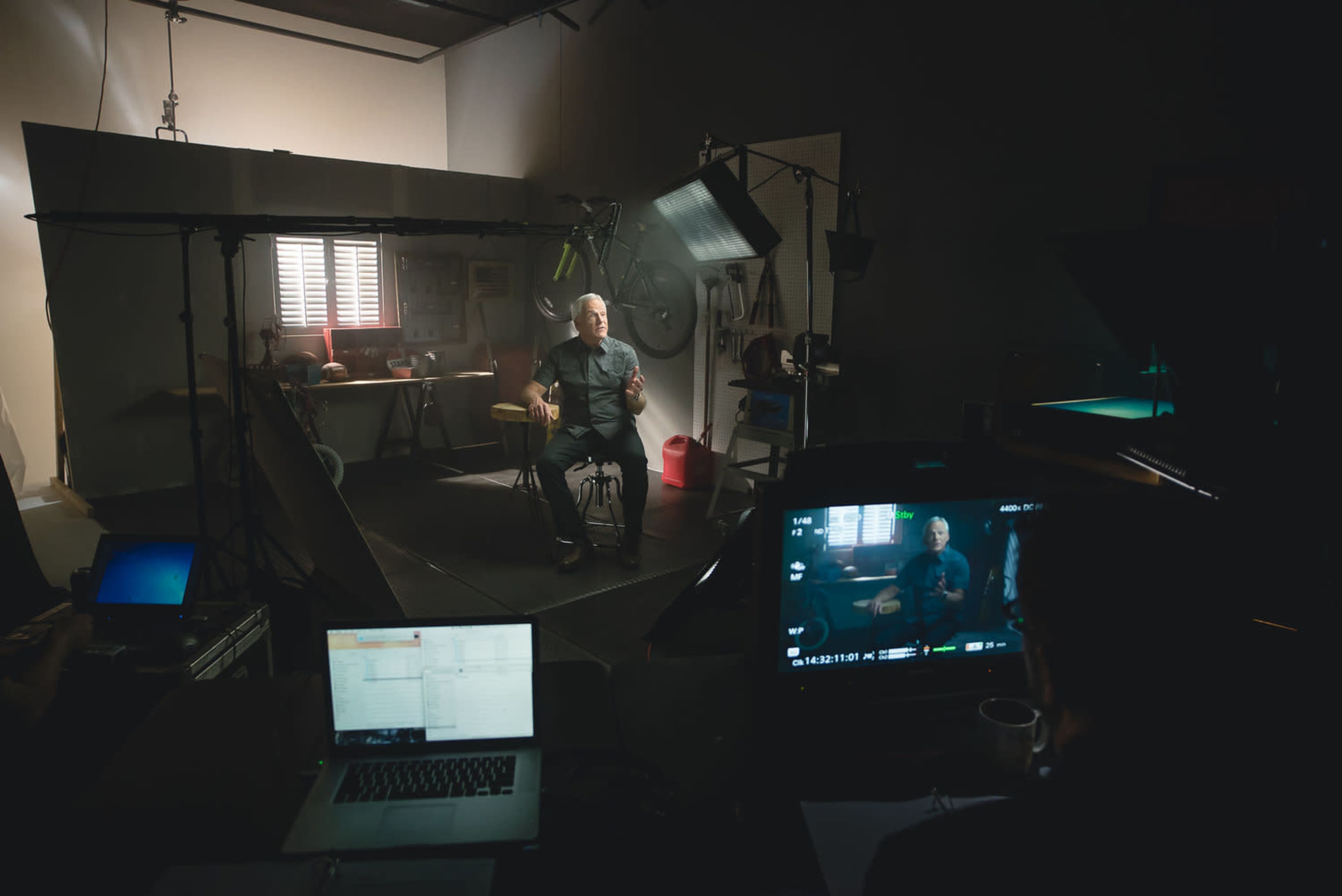 A man sits on a chair in a studio setting, speaking into a camera while a person operates equipment and monitors on a desk nearby.