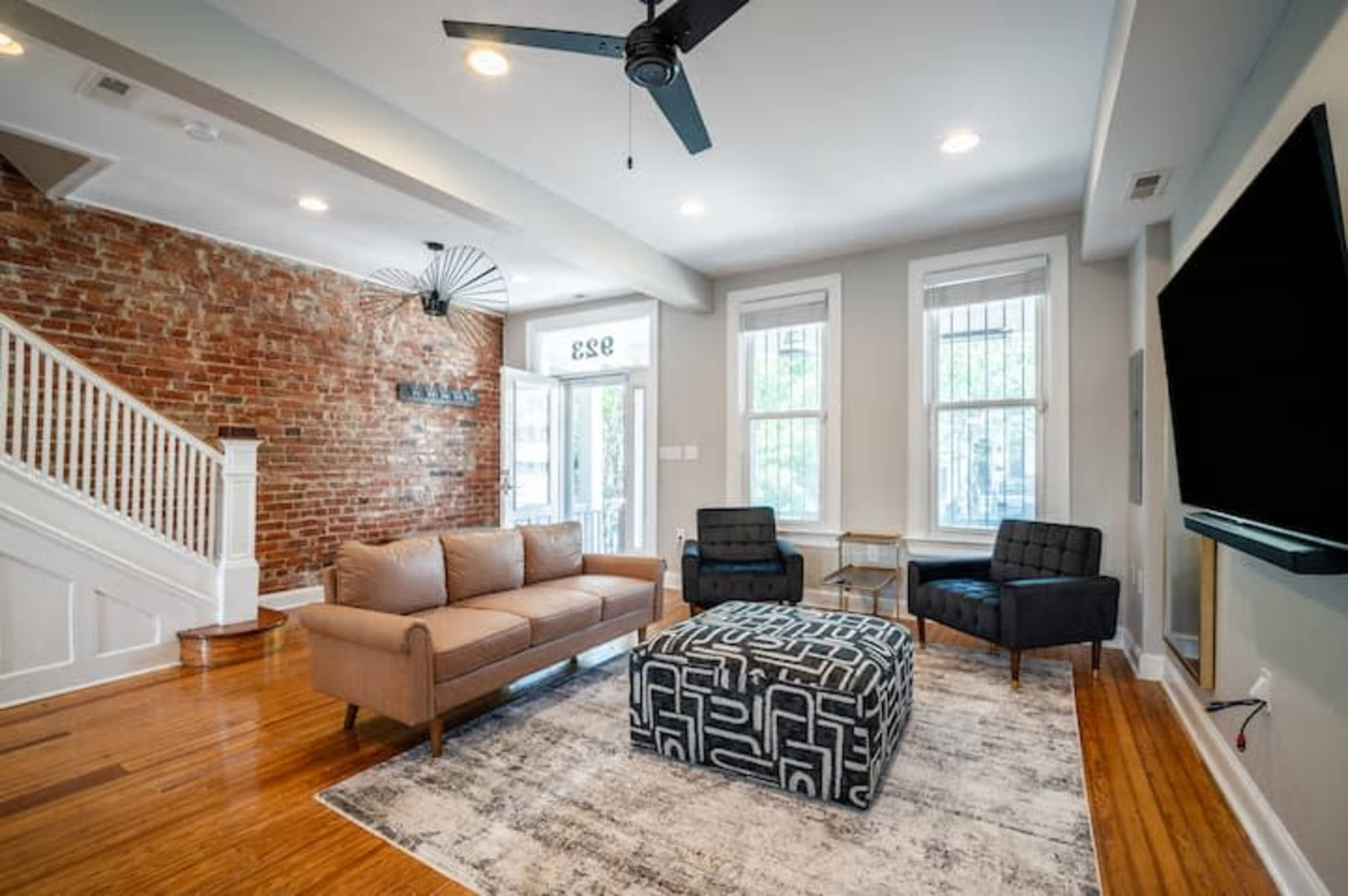 A living room featuring brick walls, a light-colored sofa, two black chairs, and a patterned ottoman on a wood floor.