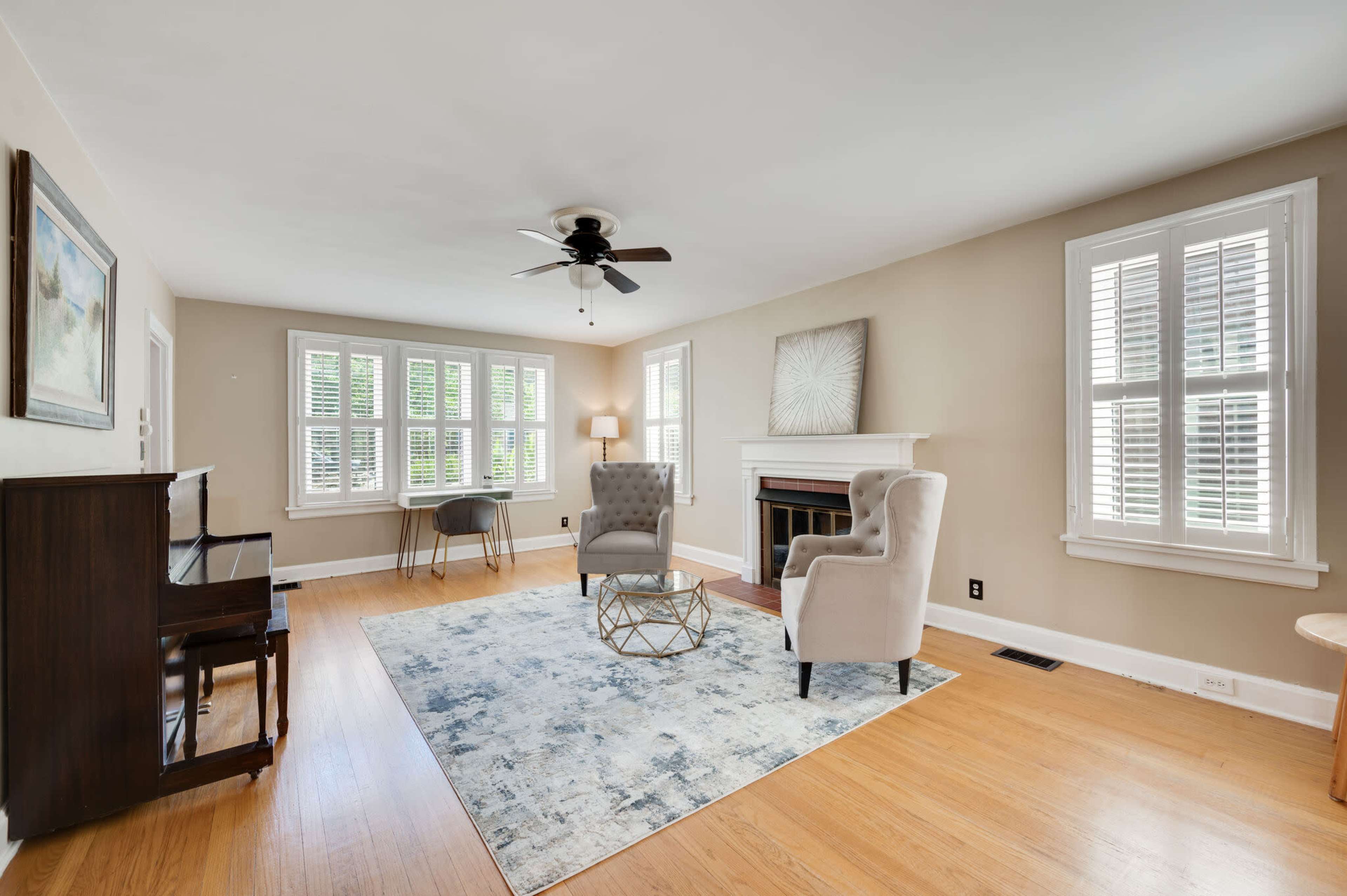 A living room features two armchairs, a coffee table, a piano, and a fireplace, with large windows allowing natural light.