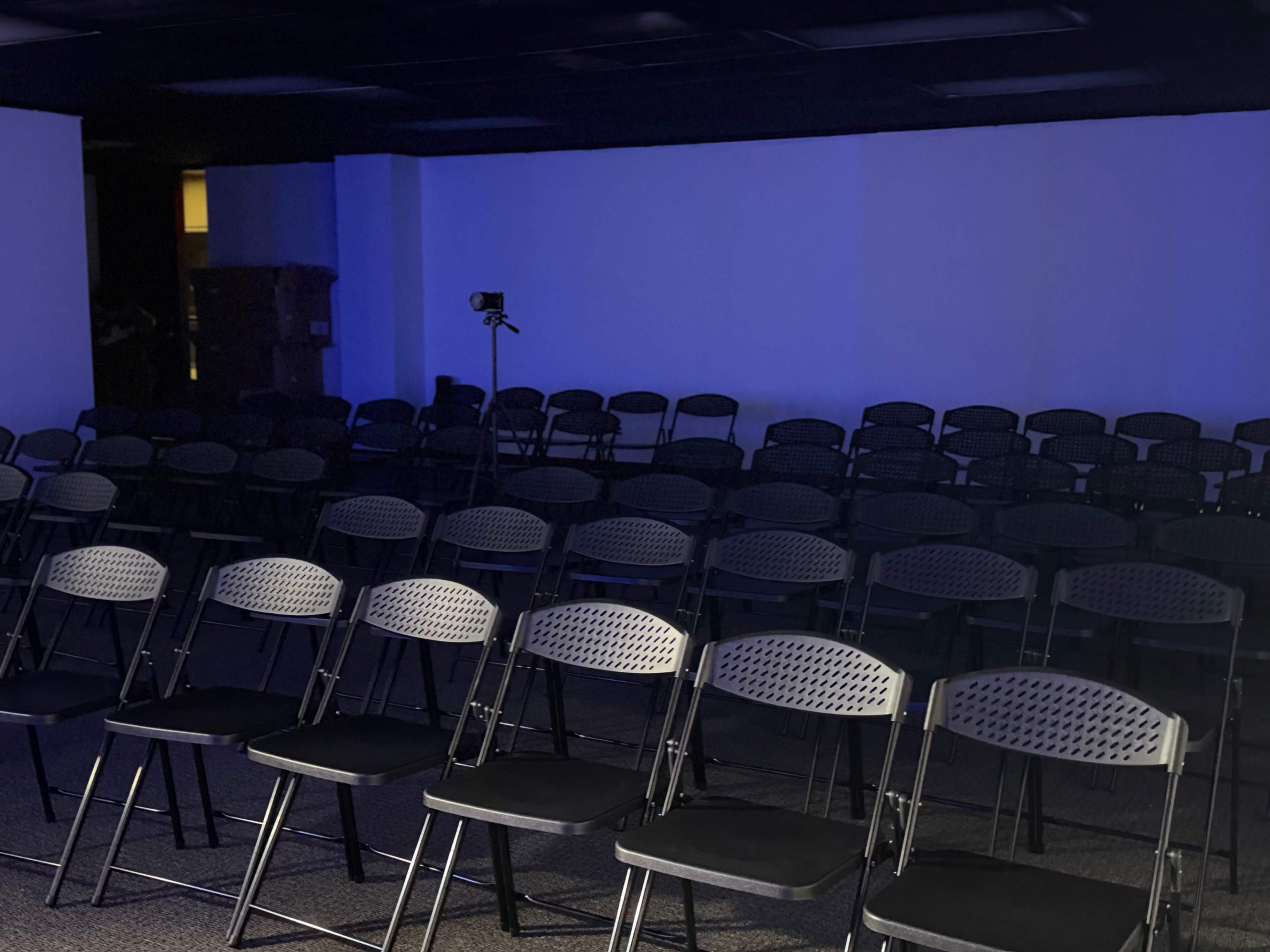 The image shows a row of black folding chairs arranged in a dimly lit room with a blue hue and a camera set up in the background.
