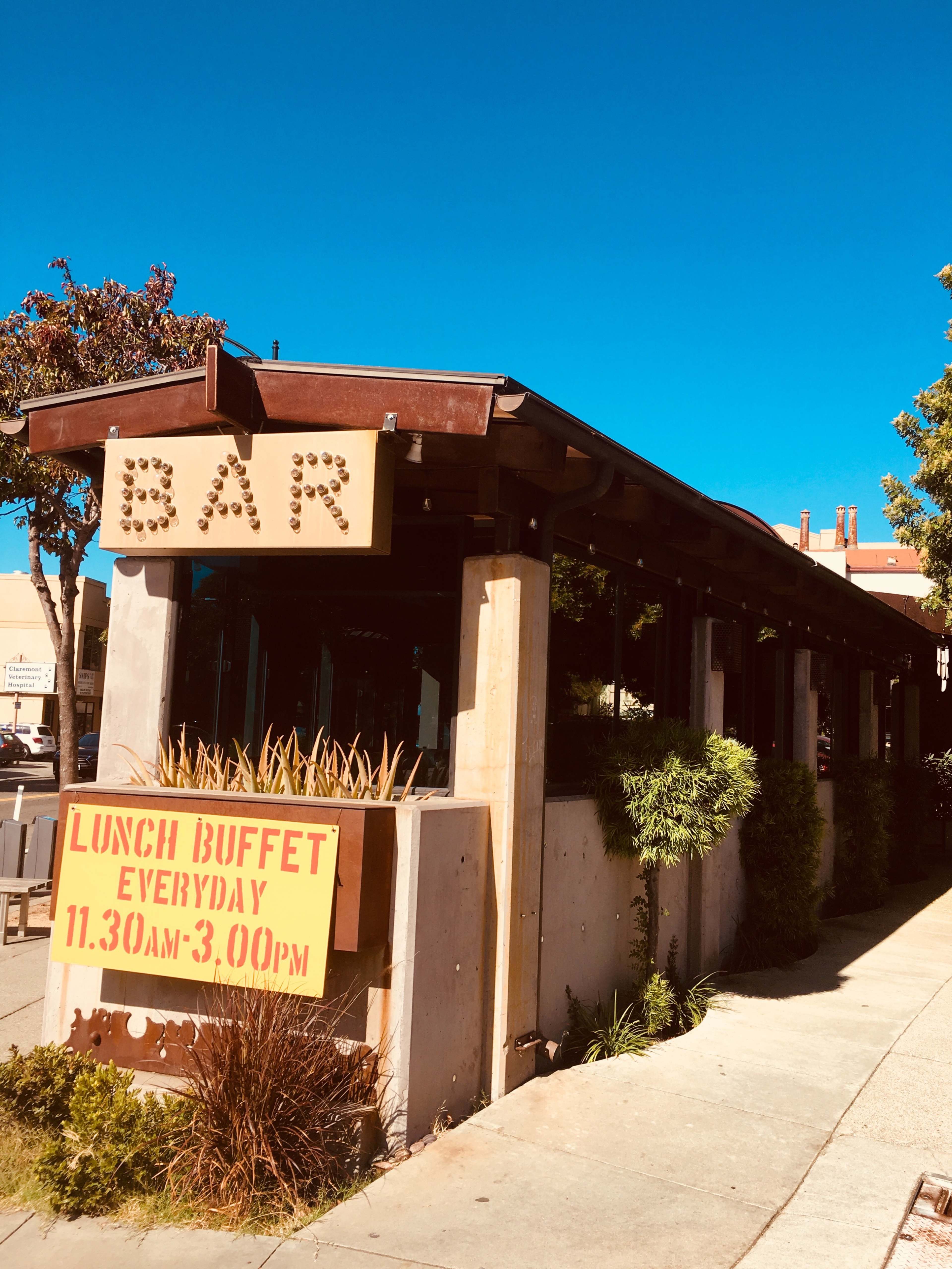 A building with a large sign reading "BAR" and a poster advertising a lunch buffet is located on a clear day with a blue sky.
