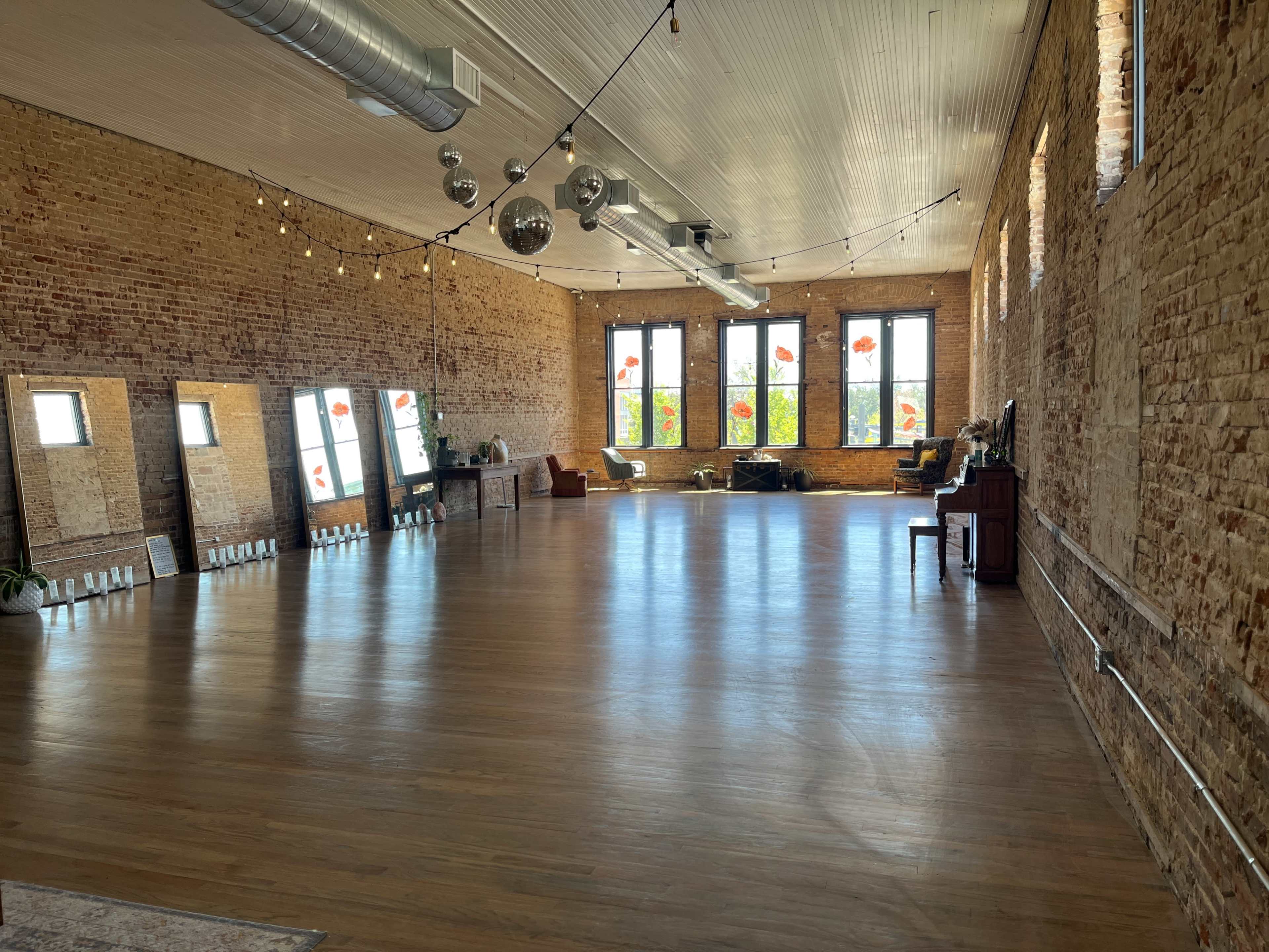 The image shows a spacious, empty room with brick walls, large windows, and wooden flooring illuminated by overhead lights and disco balls.