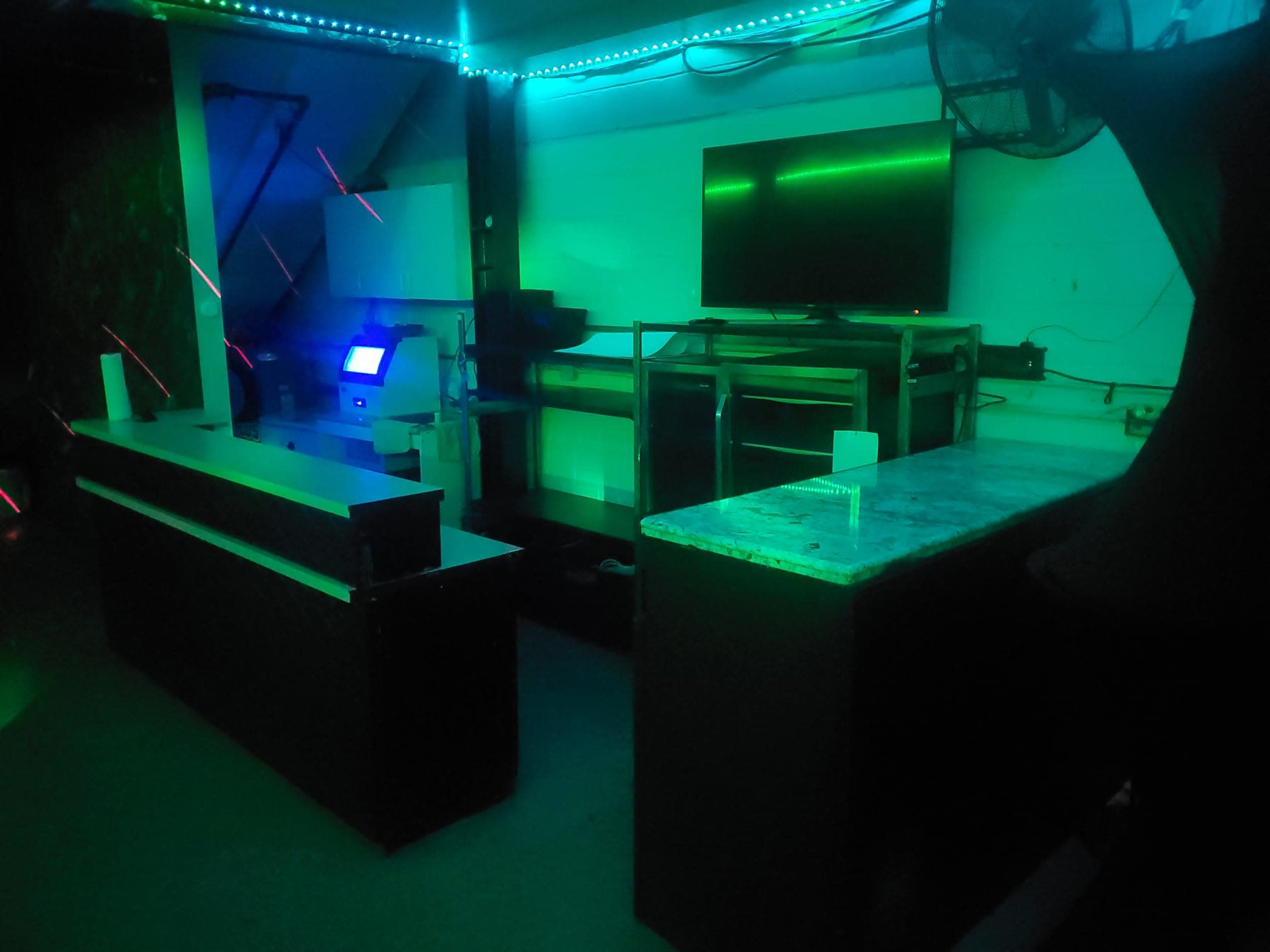 The image shows a dimly lit room featuring a bar area with two countertops, a large television screen, and ambient green lighting.