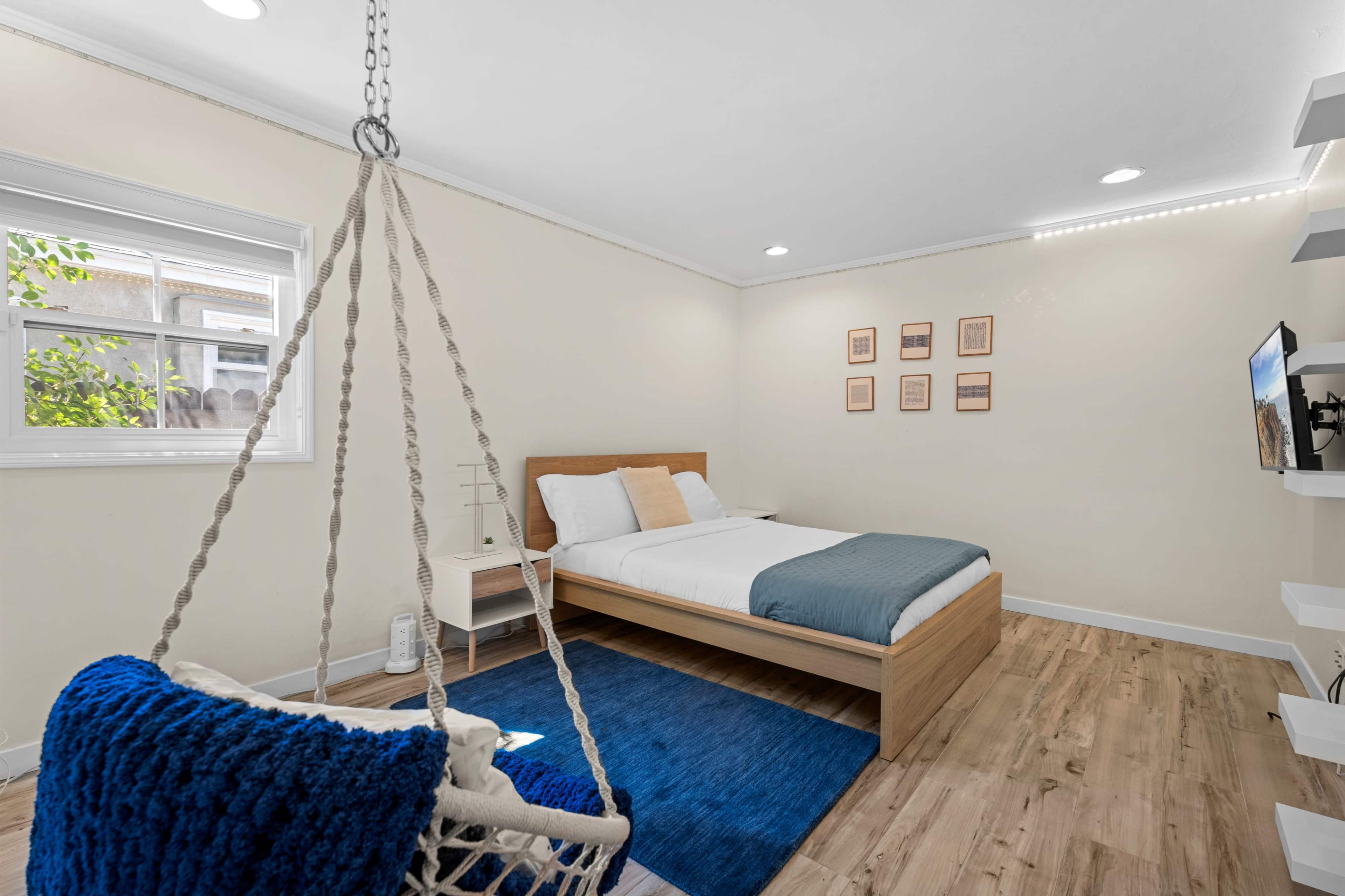 The image shows a minimalist bedroom featuring a double bed, a hanging chair, and a wall-mounted TV, with light wood flooring and neutral-colored walls.