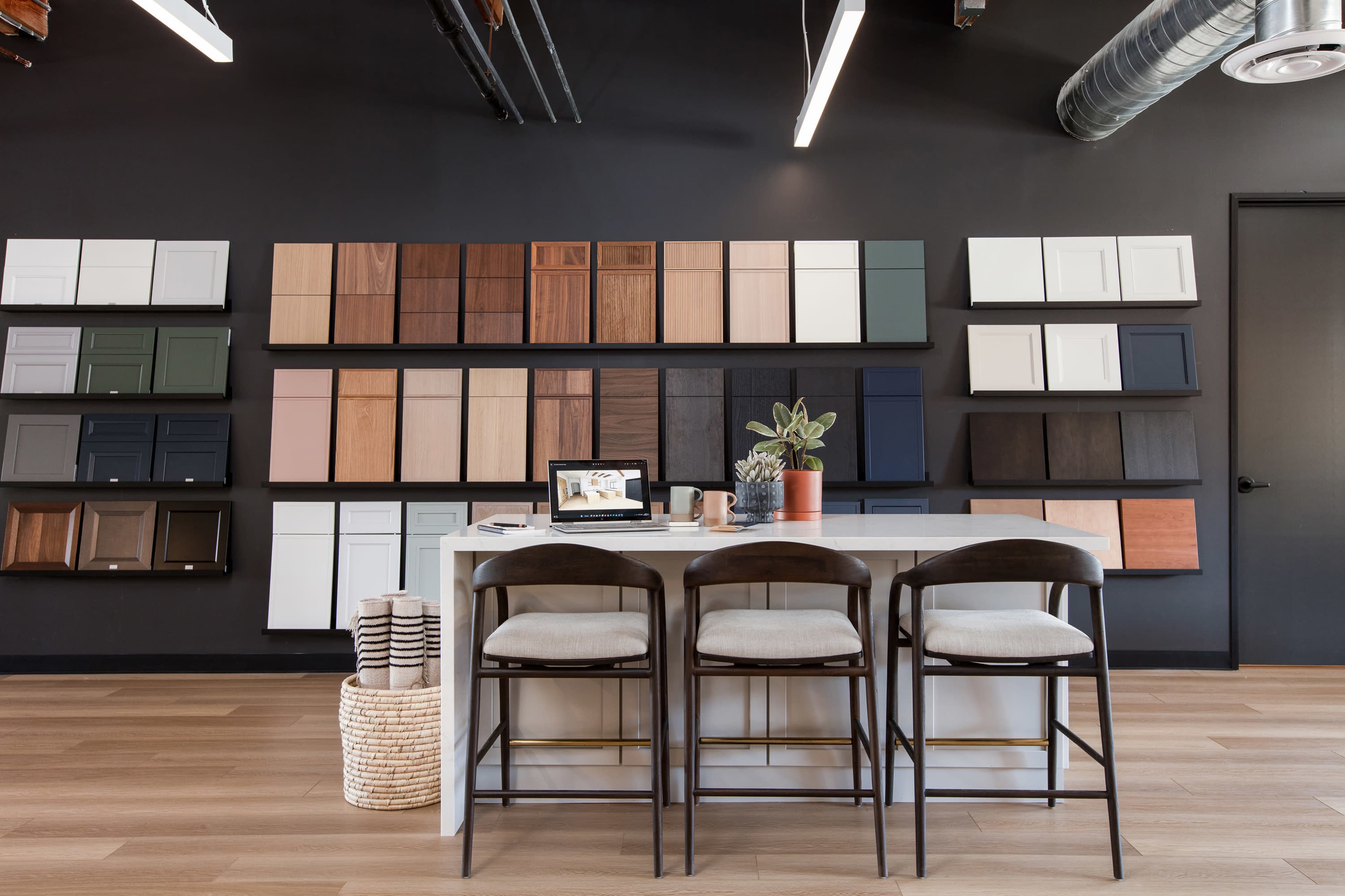 A modern workspace features a table with three bar stools and a selection of cabinet samples displayed on the wall behind it.