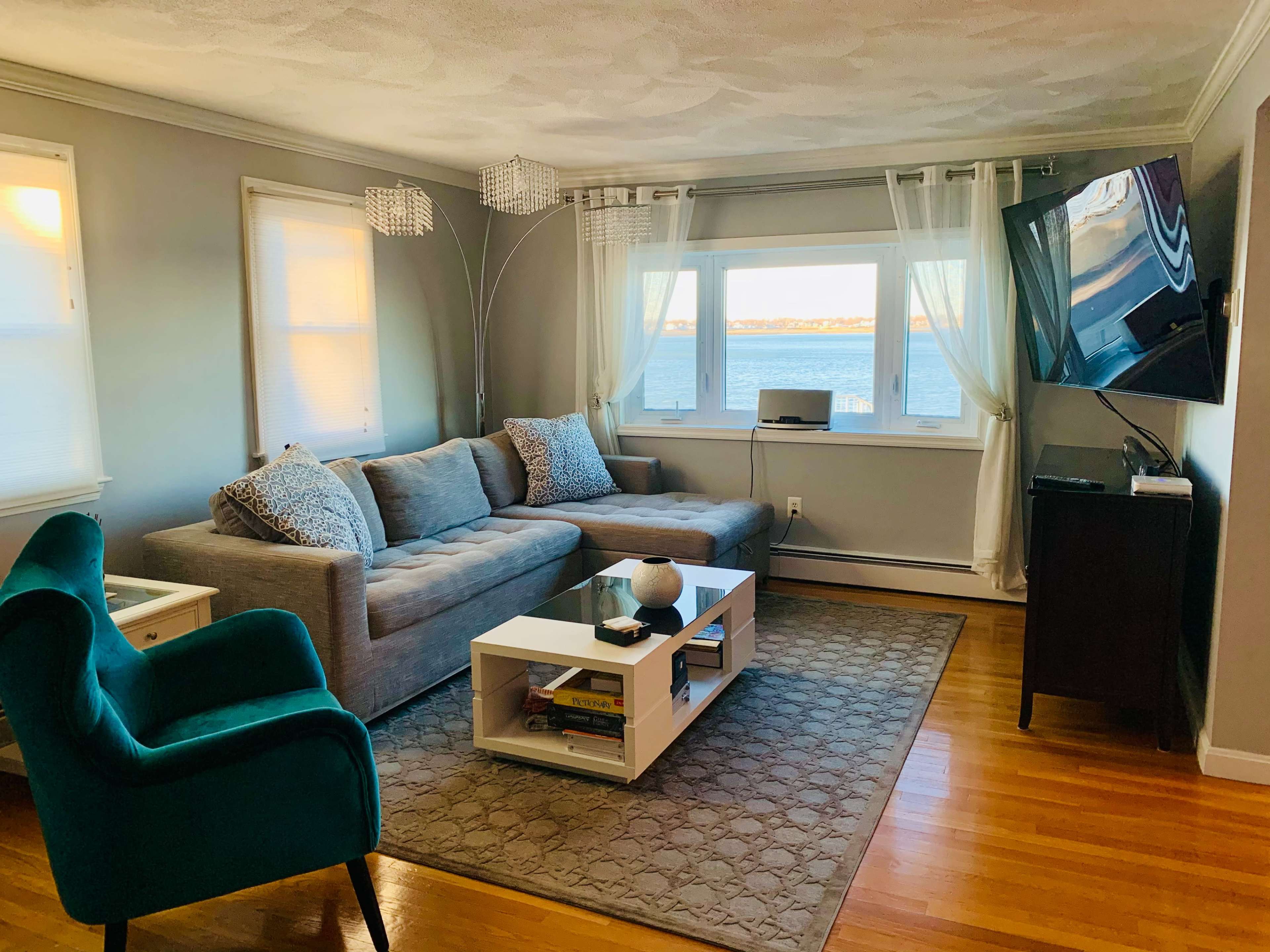 The living room features a gray sofa and an armchair, with a coffee table and a television, overlooking a water view through large windows.