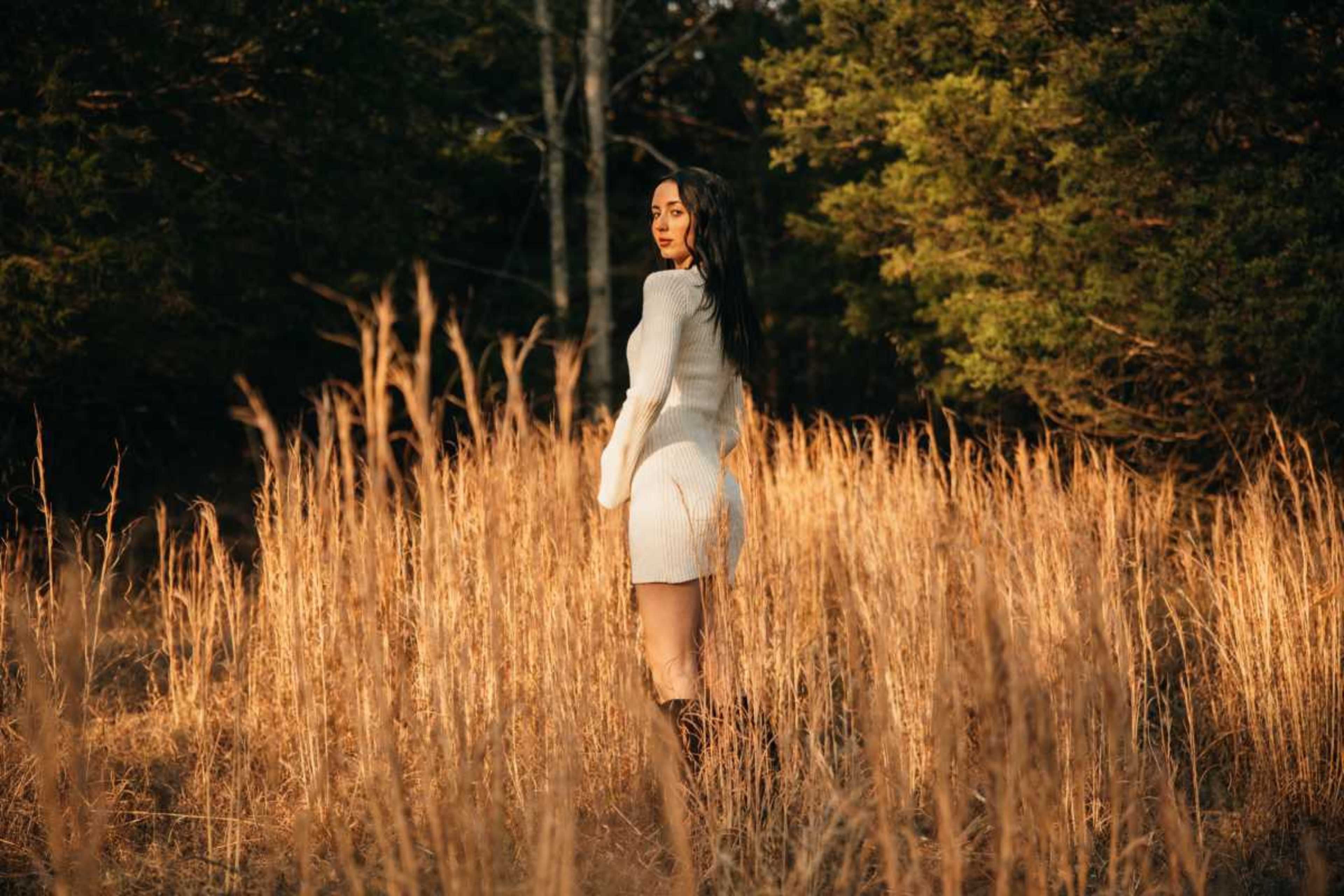 A woman in a white dress stands in a field of tall, golden grass with trees in the background during sunset.