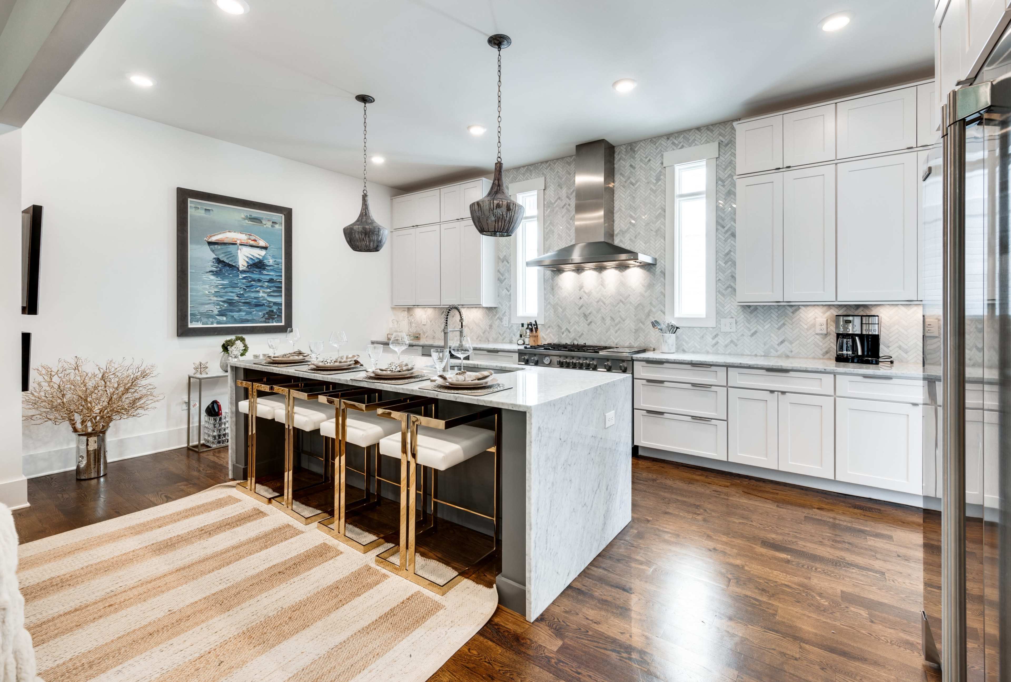 The image shows a modern kitchen featuring white cabinetry, a marble island with gold-accented bar stools, and large windows allowing natural light.