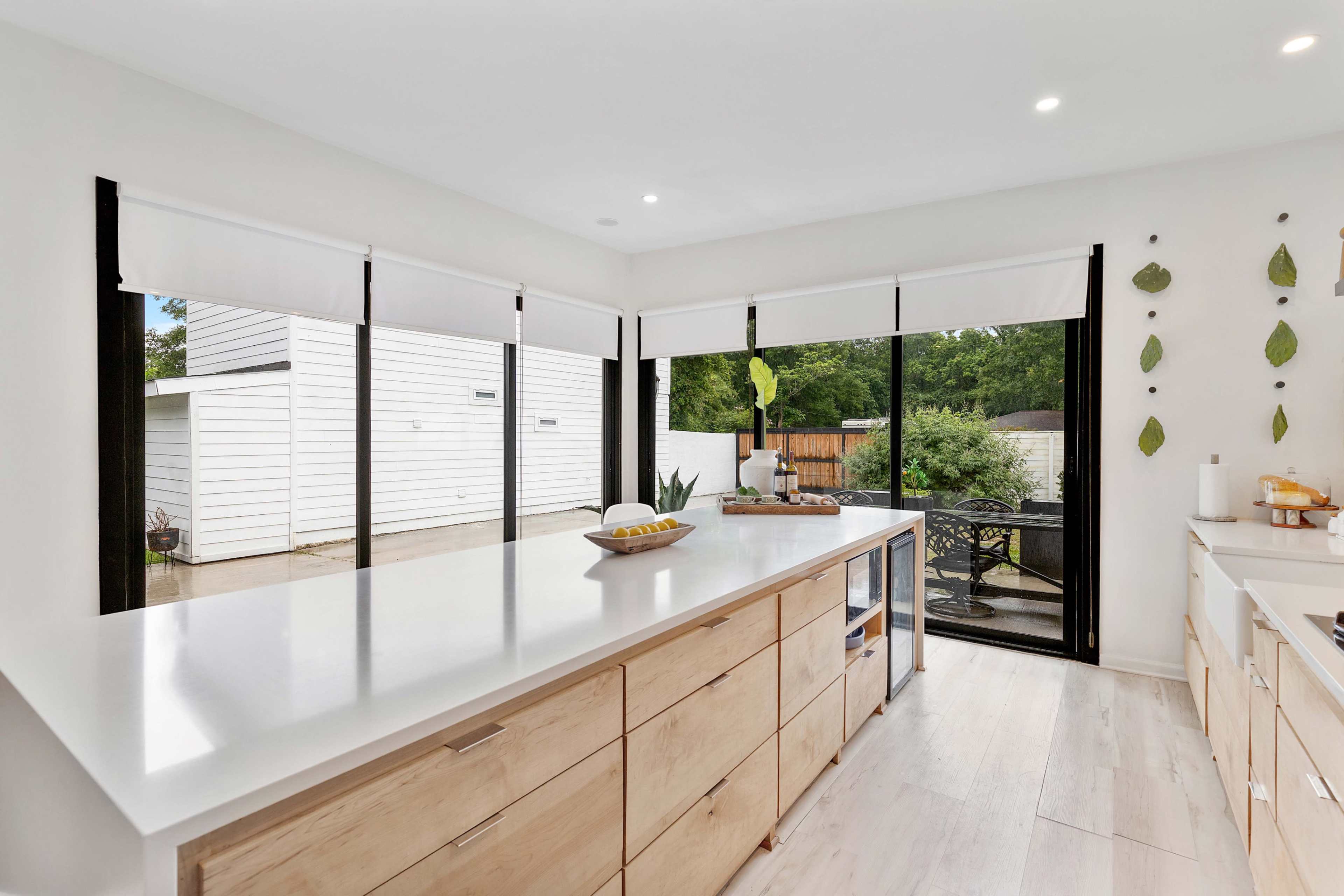 The image shows a modern kitchen with an island, large glass windows overlooking an outdoor patio, and minimalistic cabinetry.