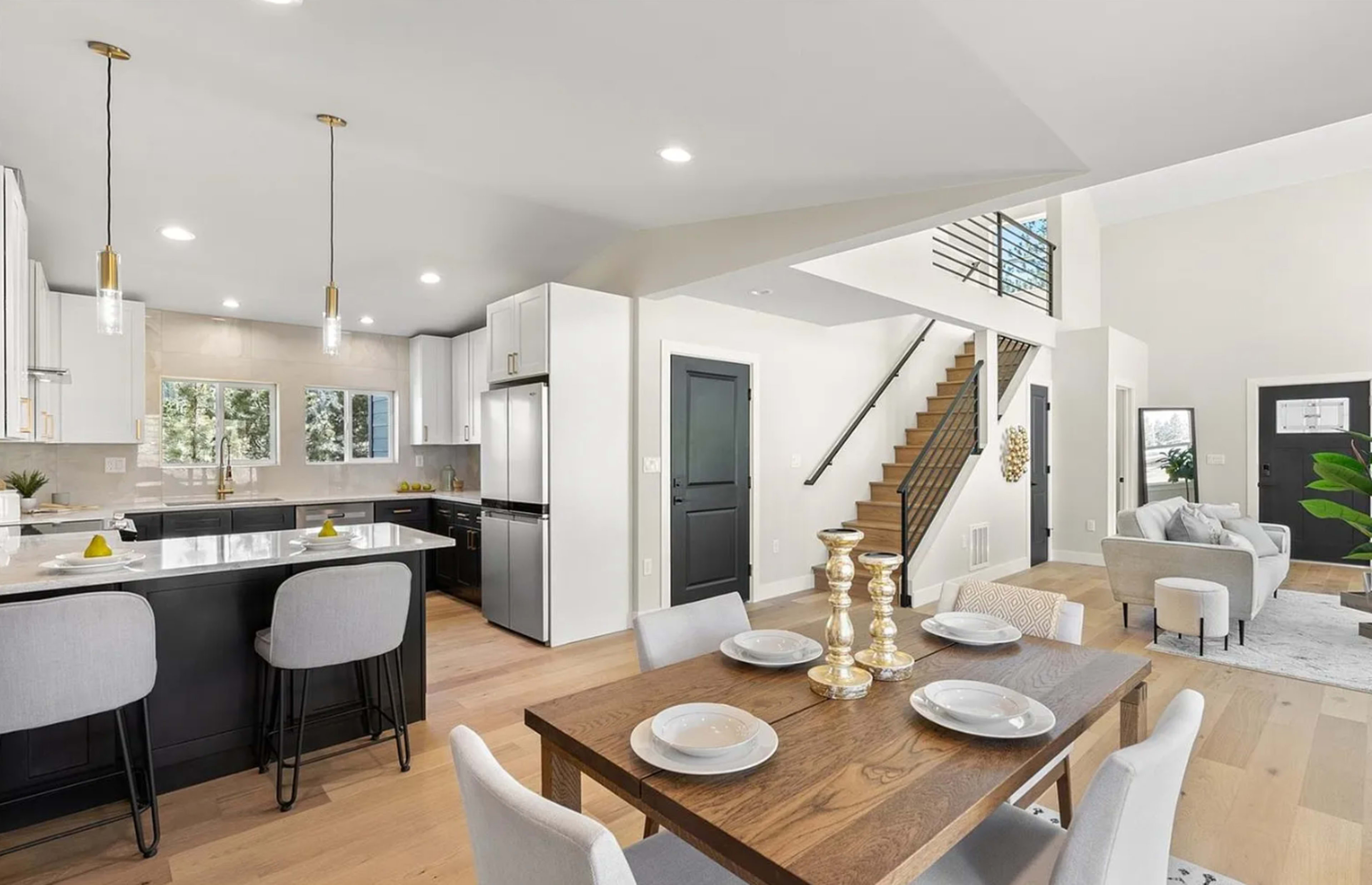 The image features a modern kitchen adjacent to a dining area, showcasing a wooden dining table set for four, with sleek countertops and a staircase leading to an upper level in the background.