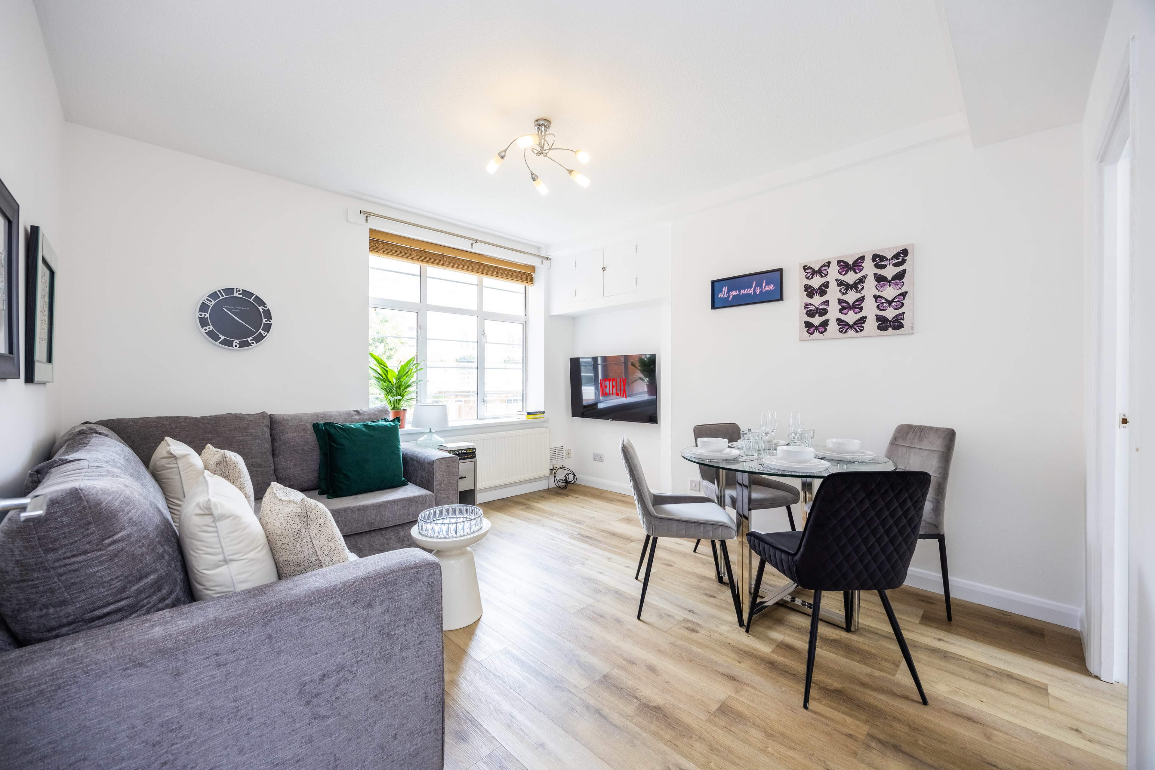 A cozy living area features a gray sofa, a round dining table, and a television against a bright wall with decorative art and a window.