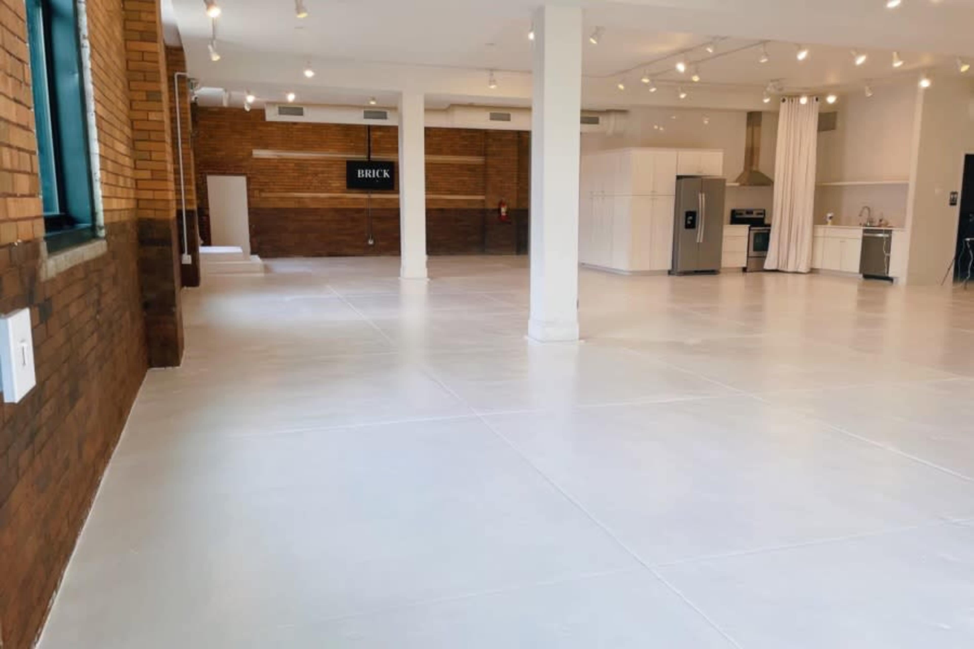 A spacious, empty room with a polished white floor, exposed brick walls, and a modern kitchen area visible in the background.