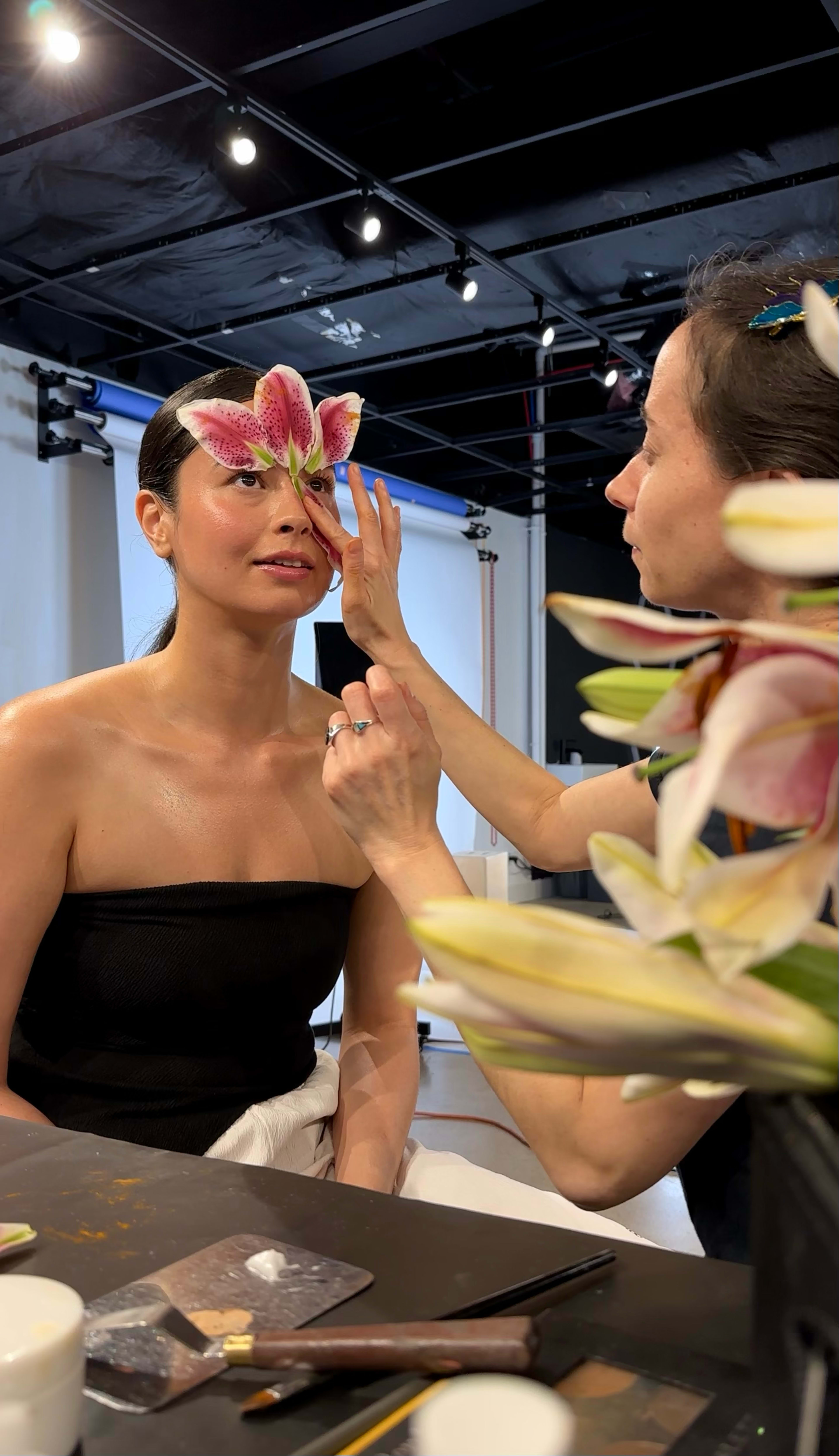 A makeup artist applies makeup to a model while a large flower is positioned on the model's forehead.