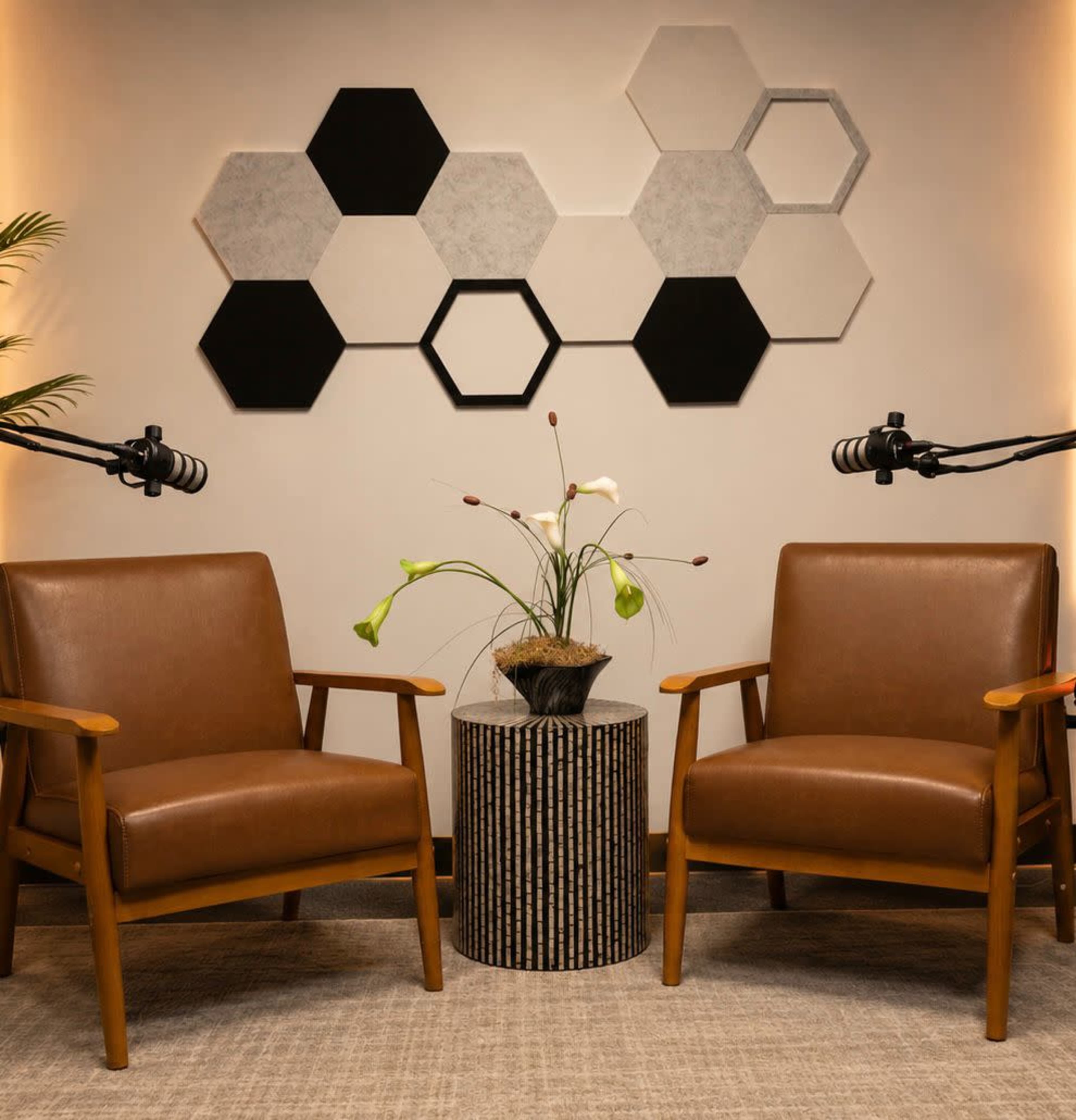 The image shows two brown wooden armchairs facing each other, with a round striped side table between them and a plant arrangement on top, against a wall decorated with hexagonal shapes in black, white, and gray.