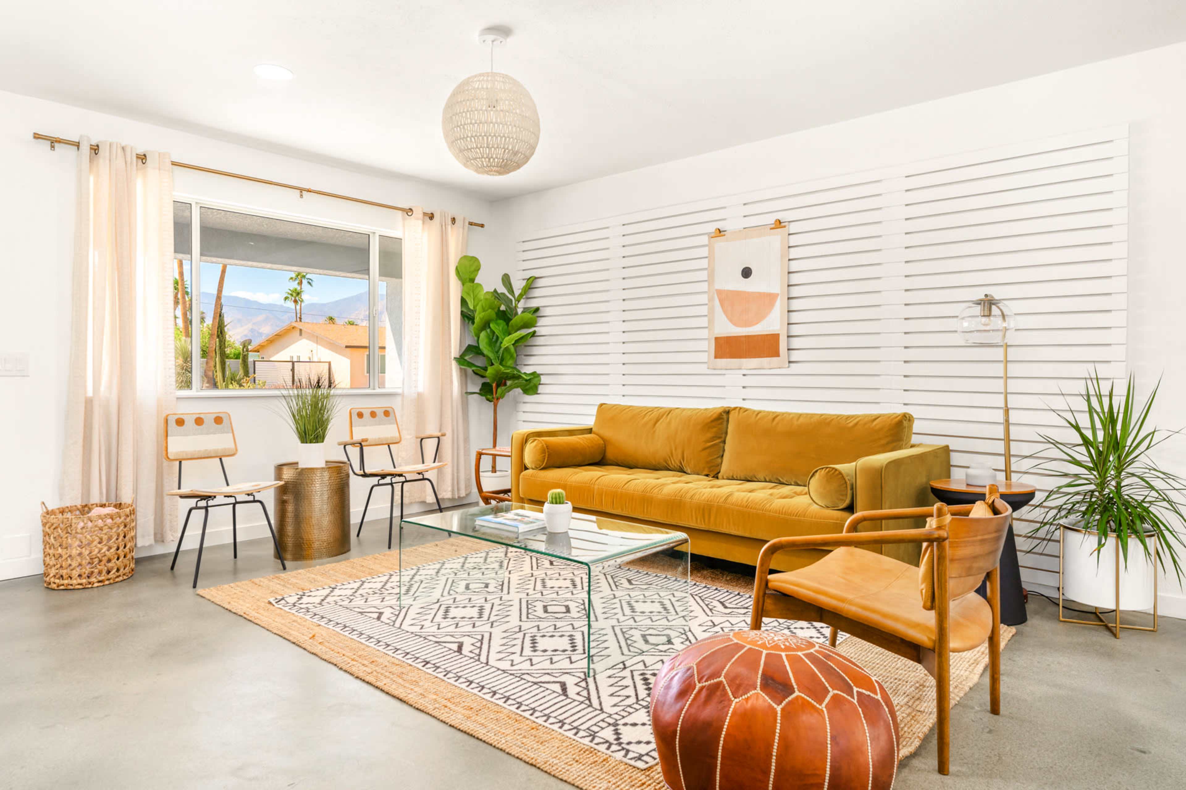 A modern living room features a mustard yellow sofa, a glass coffee table, and various plants against a backdrop of white walls and wooden slats.