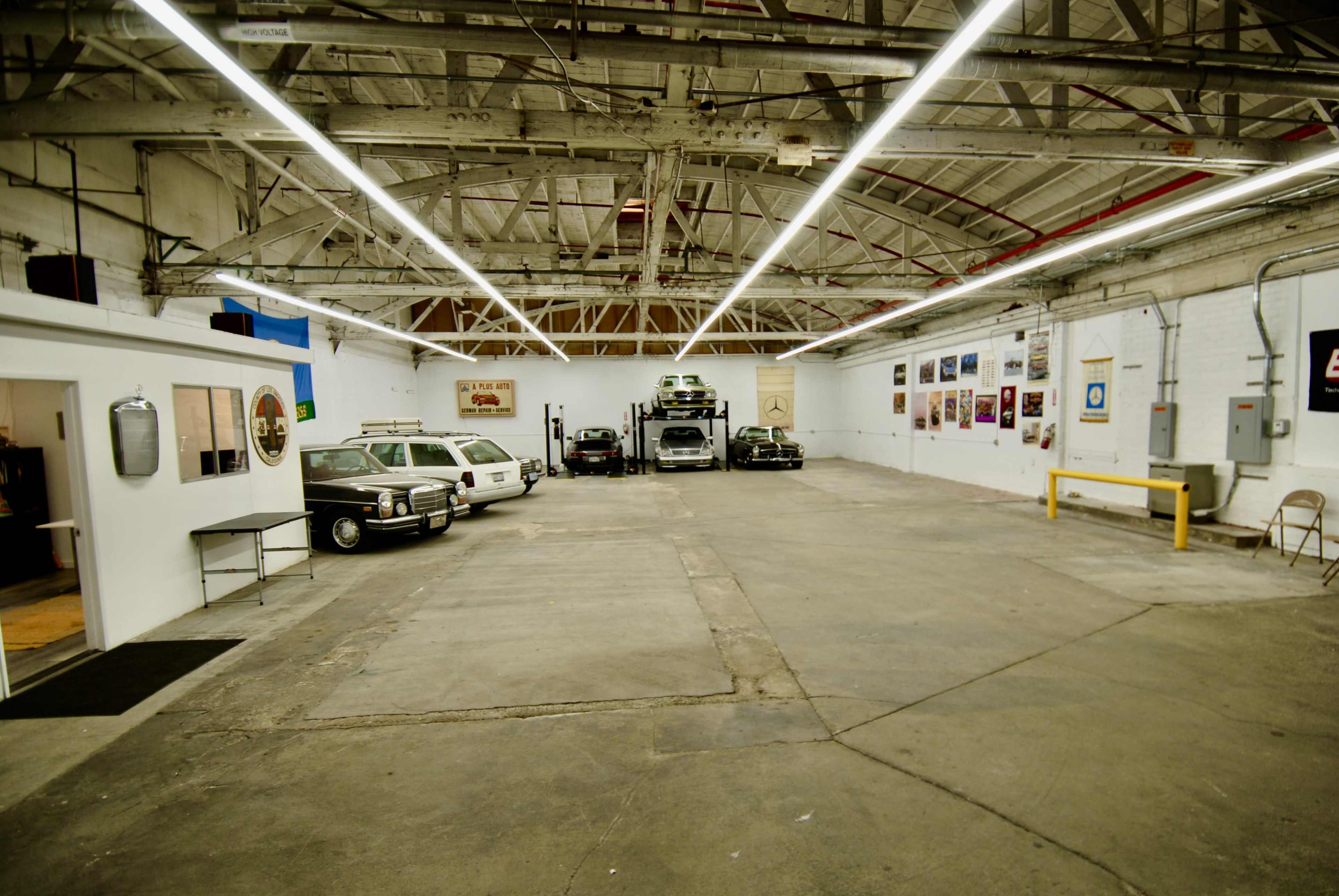 The image shows a spacious industrial garage with several vehicles, including vintage cars, parked along one side and artwork displayed on the walls.
