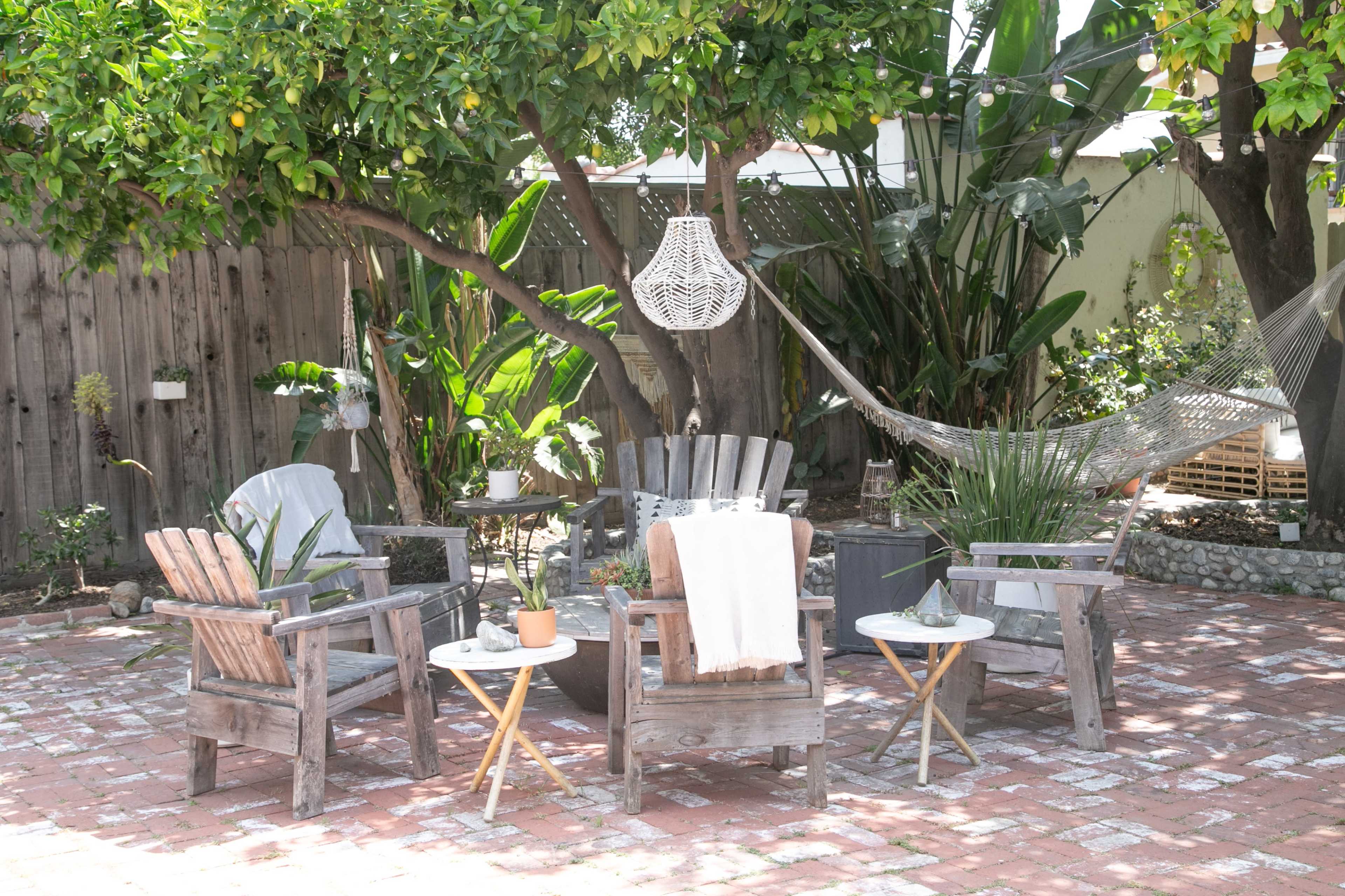 A backyard patio features wooden chairs and tables under a tree, with a hammock hanging nearby.