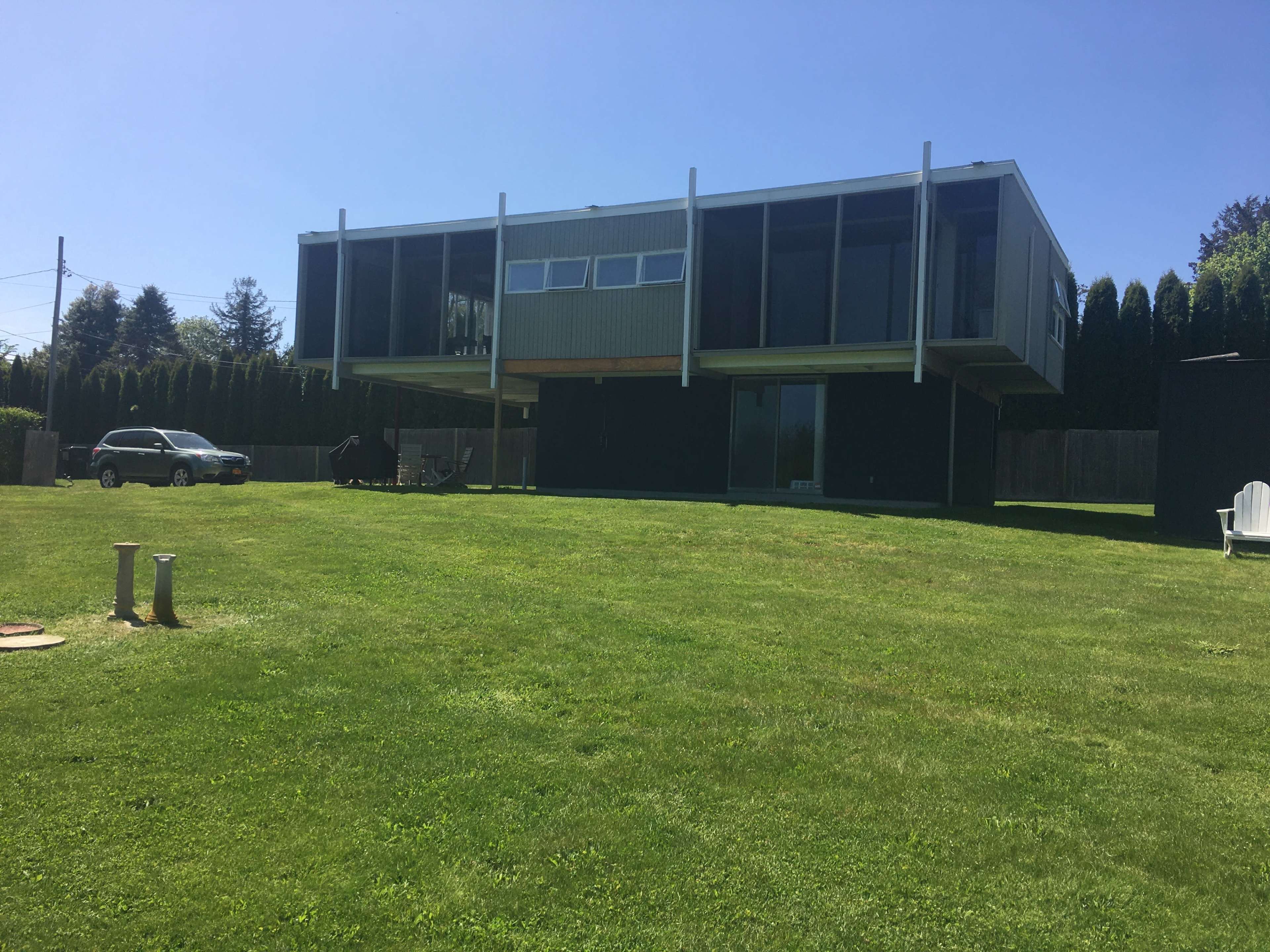 A modern house raised on stilts with large windows, surrounded by a well-maintained lawn and a parked car nearby.