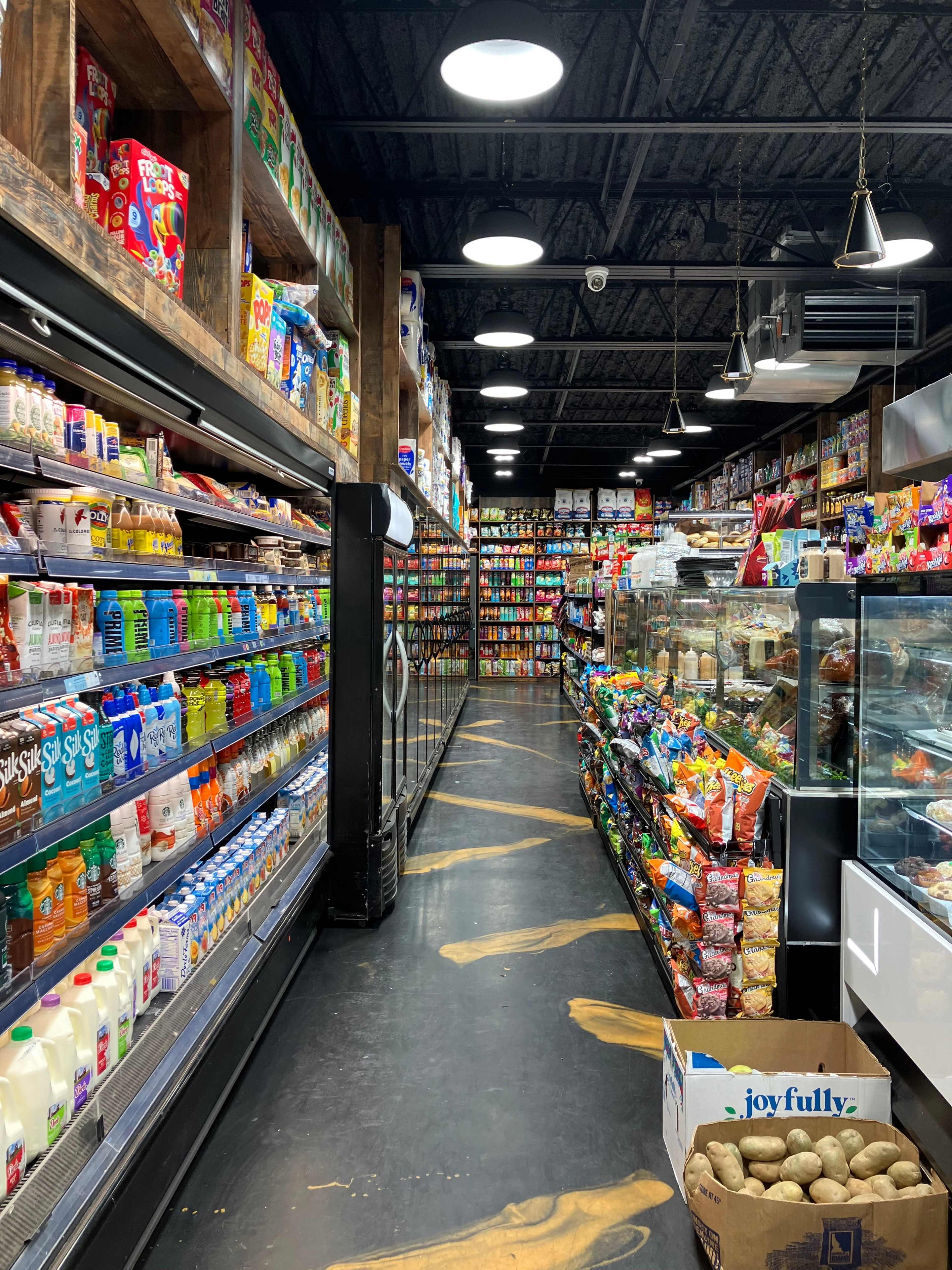 An aisle in a grocery store lined with shelves of colorful packaged goods, dairy products, and snacks, with a display case of food on one side.