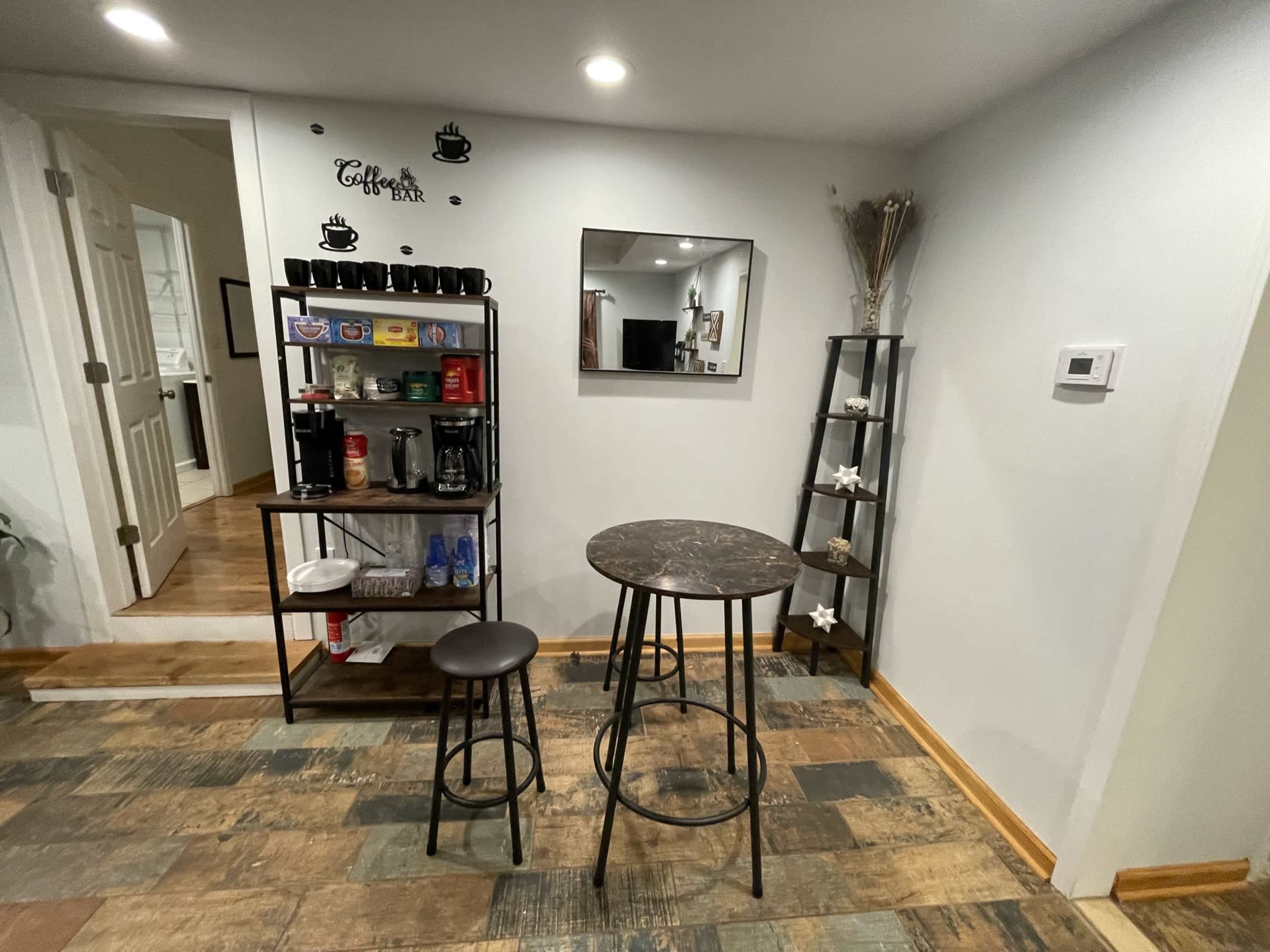 A kitchenette area features a coffee station with an assortment of mugs, a small table with a stool, and a mirror on the wall.