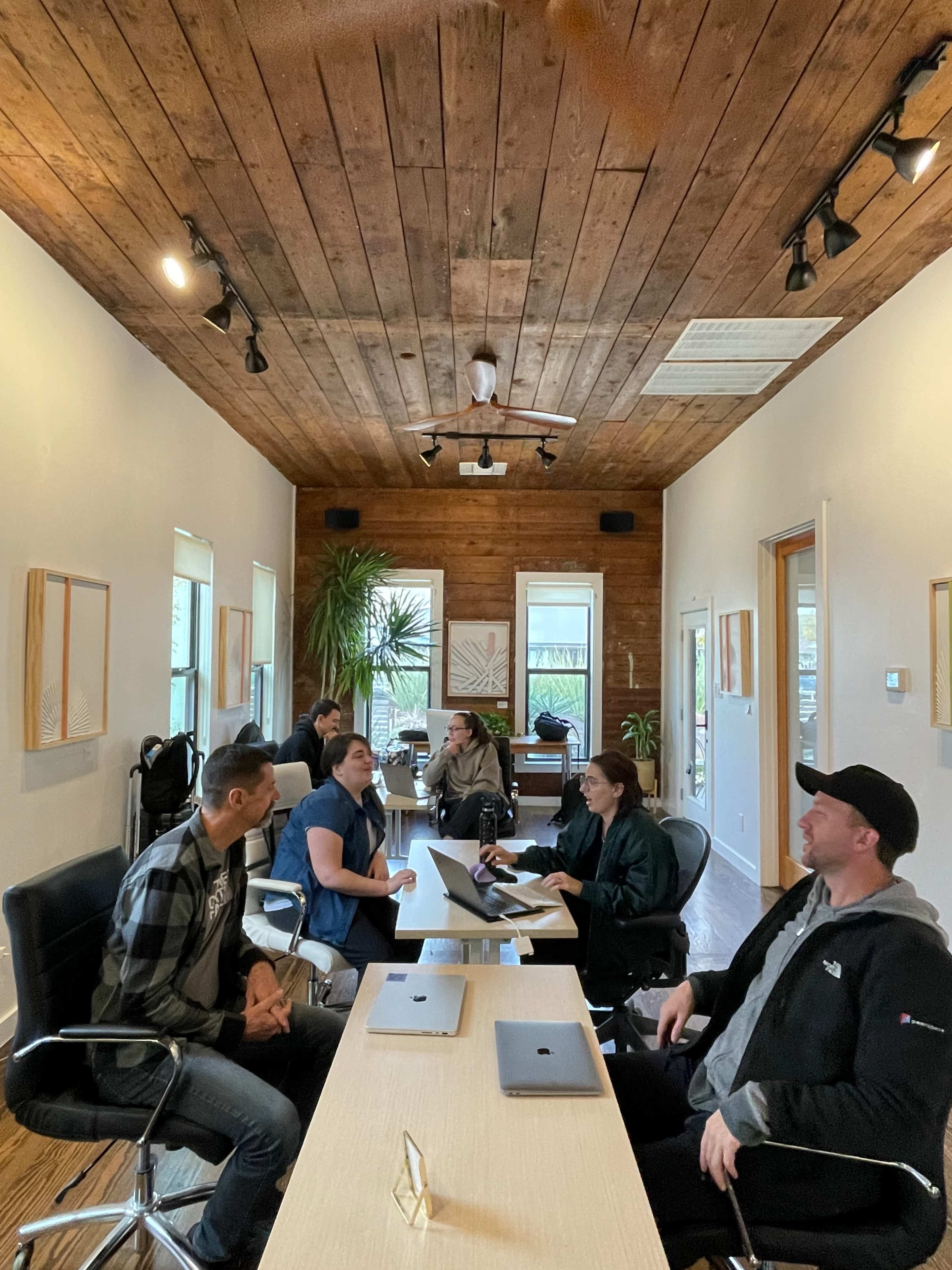 A group of six people engage in a discussion around a long table in a modern office space with wooden accents.