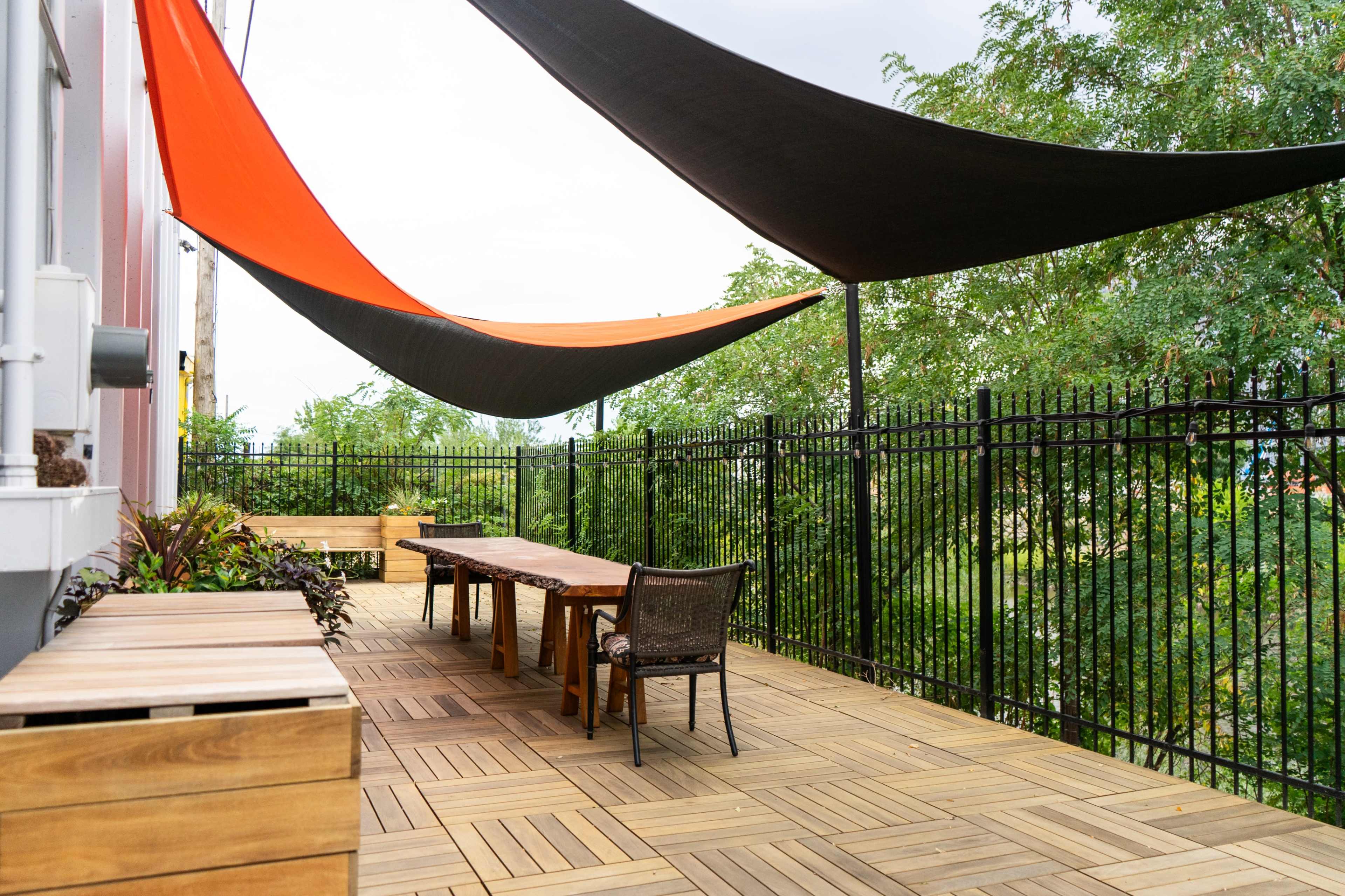 A wooden terrace features two long tables and chairs beneath decorative shade sails in orange and black, surrounded by greenery.