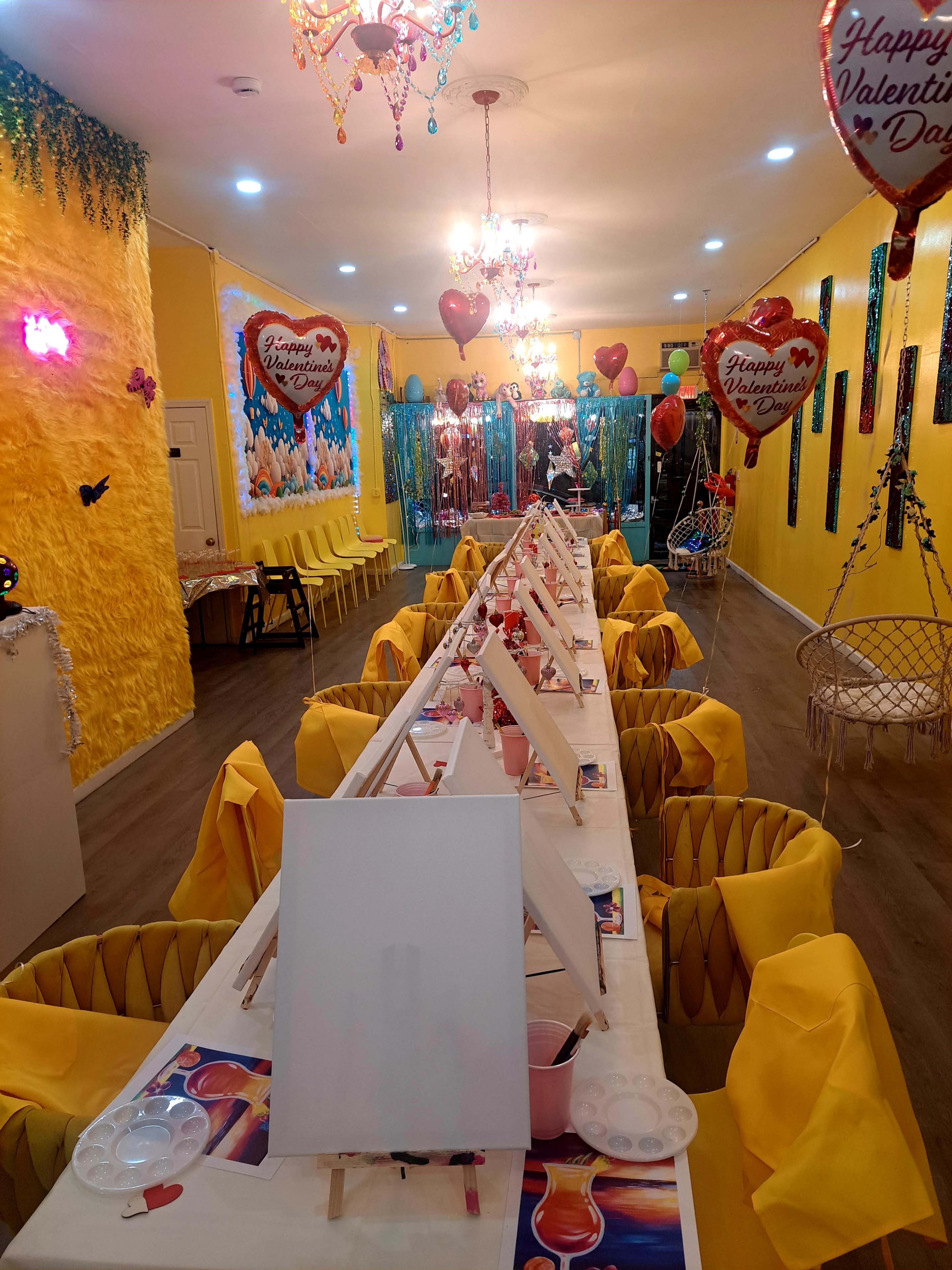 A brightly decorated room is set up for a Valentine's Day painting event, featuring yellow walls, a long table with blank canvases, and heart-shaped balloons.