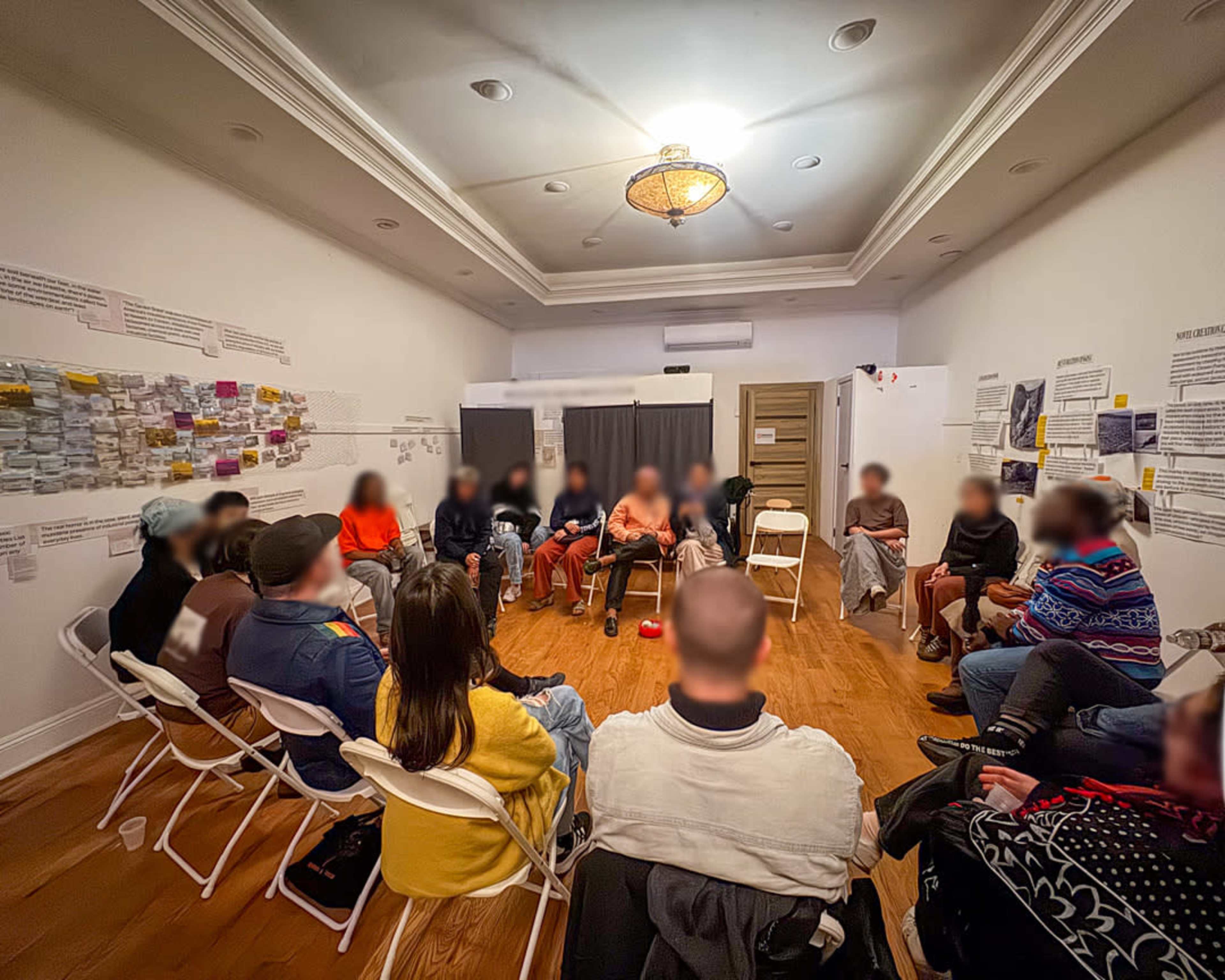 A group of attendees is seated in a circle in a well-lit room decorated with papers and notes on the walls.