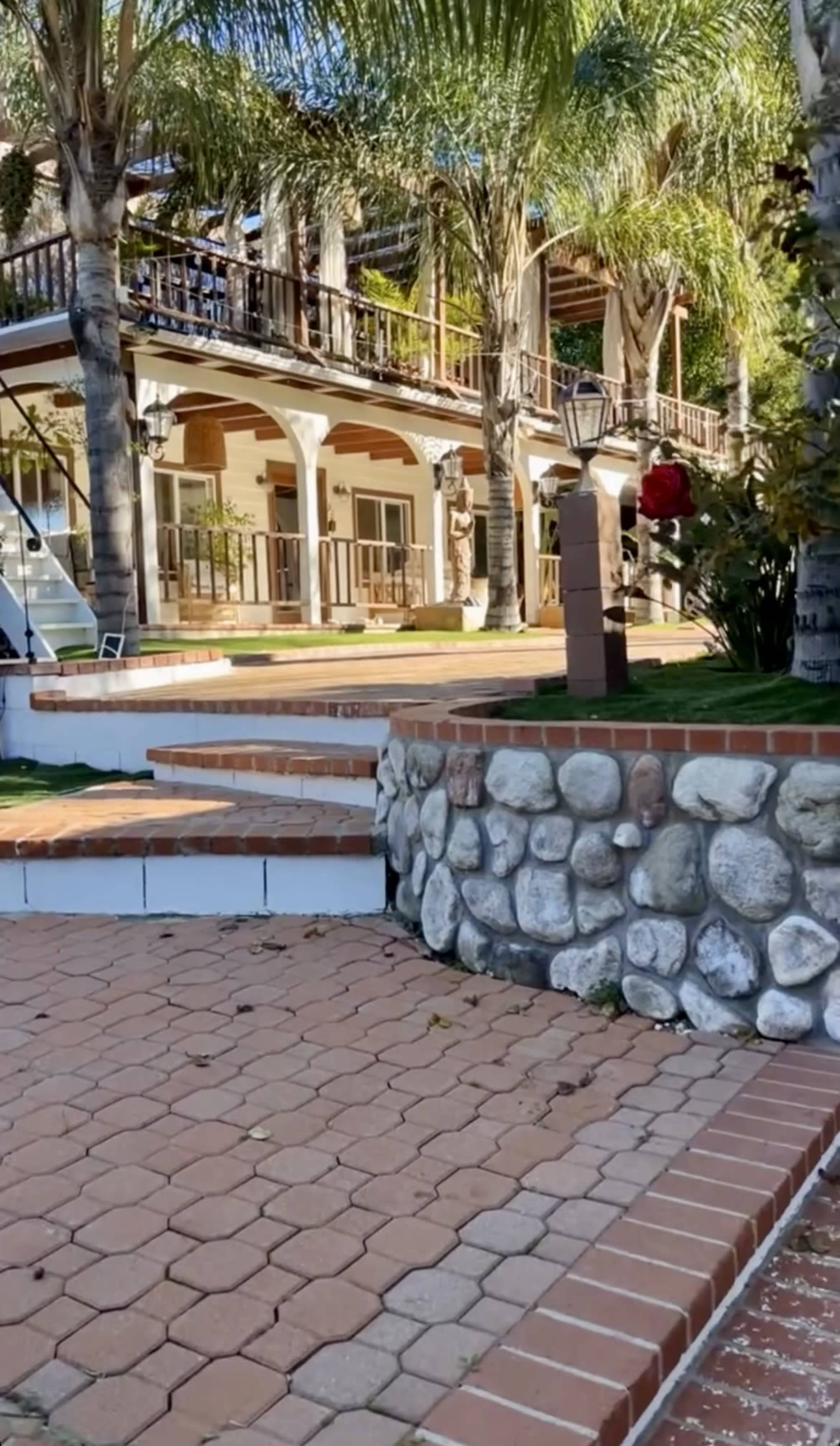The image shows a multi-level house with a porch and balconies, surrounded by palm trees and a landscaped courtyard featuring brick pathways and a stone wall.