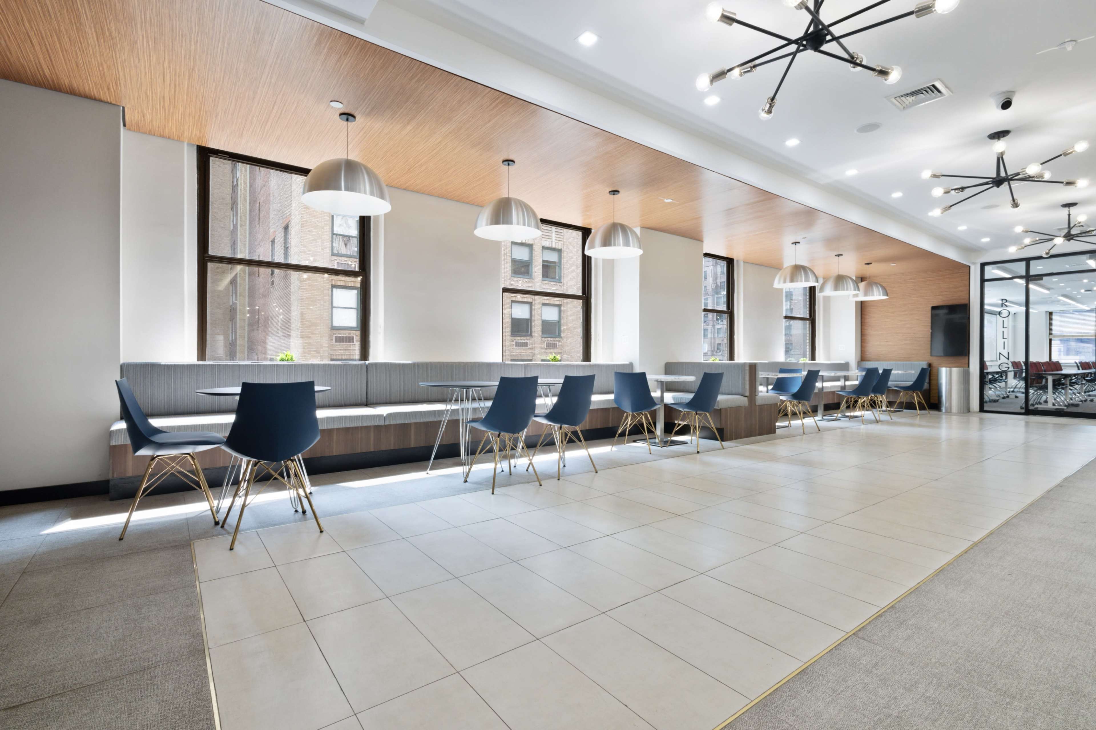 The image shows a modern indoor dining area featuring long communal tables with blue chairs, large windows, and pendant lighting.
