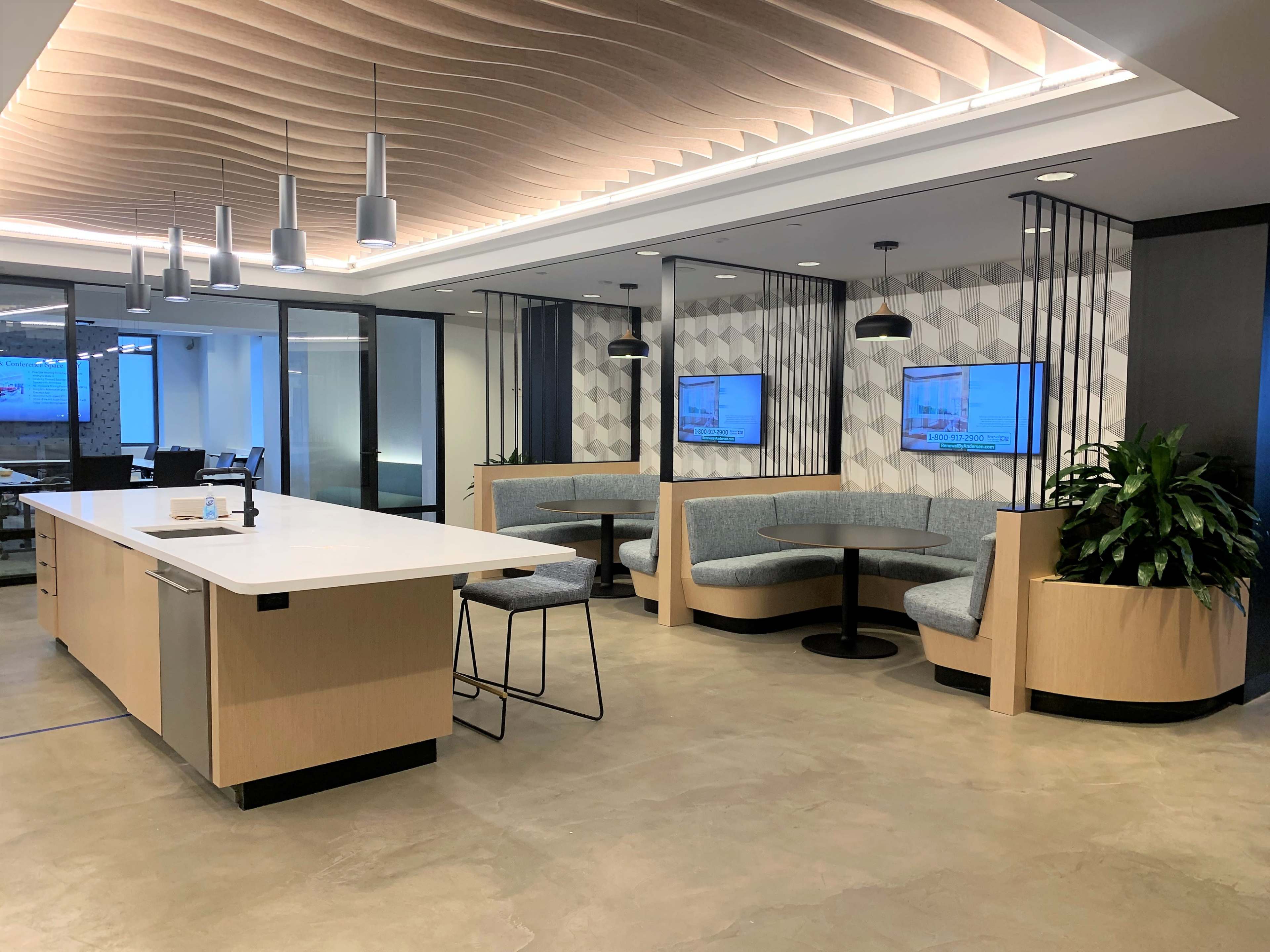 The image shows a modern office lobby area with a kitchen island, seating booths, and two televisions mounted on the walls.
