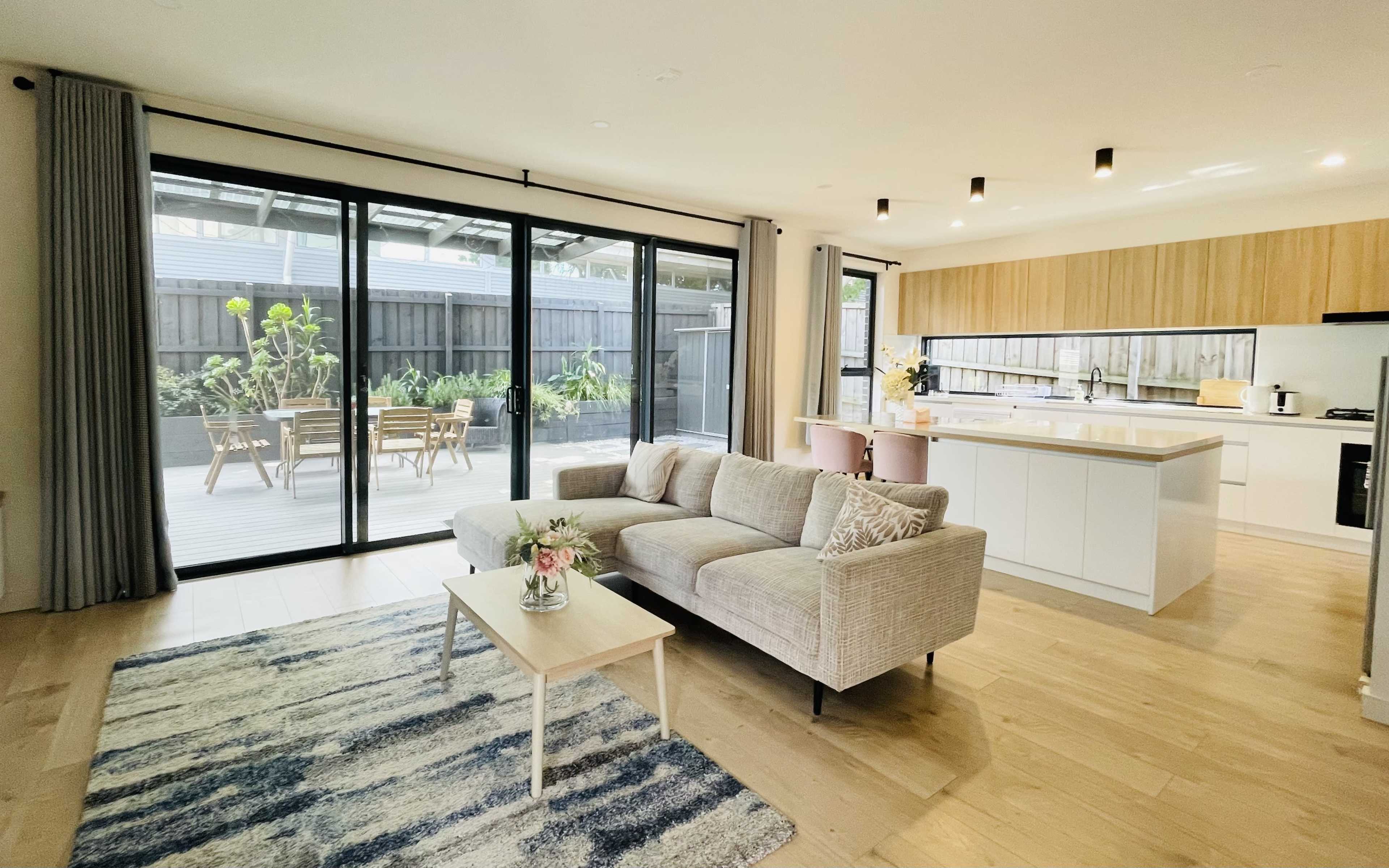A modern living room with a light-colored sofa, coffee table, and a large sliding glass door leading to an outdoor deck area with patio furniture.