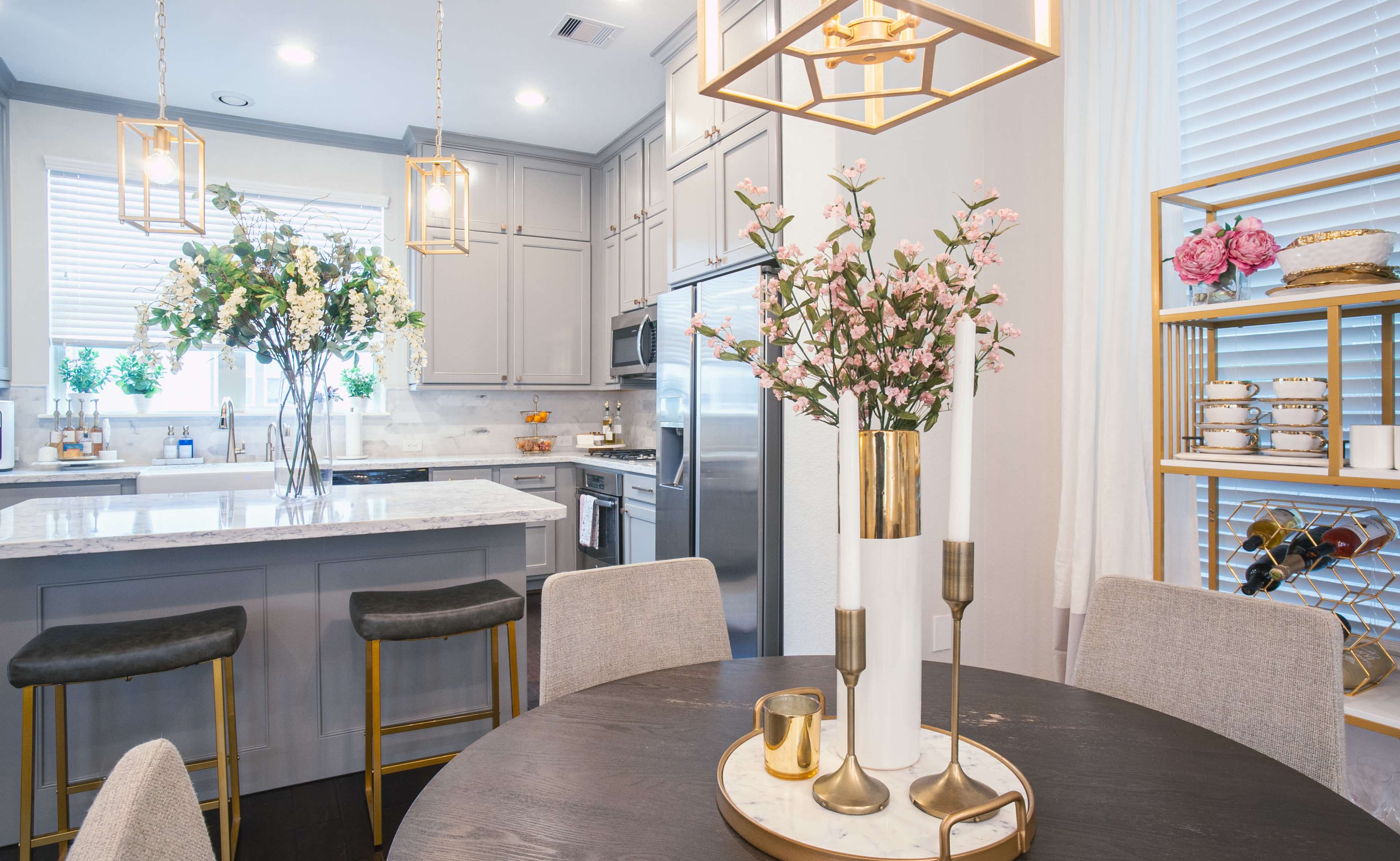 A modern kitchen features a central island with pendant lights, bar stools, and a dining table with a floral centerpiece.