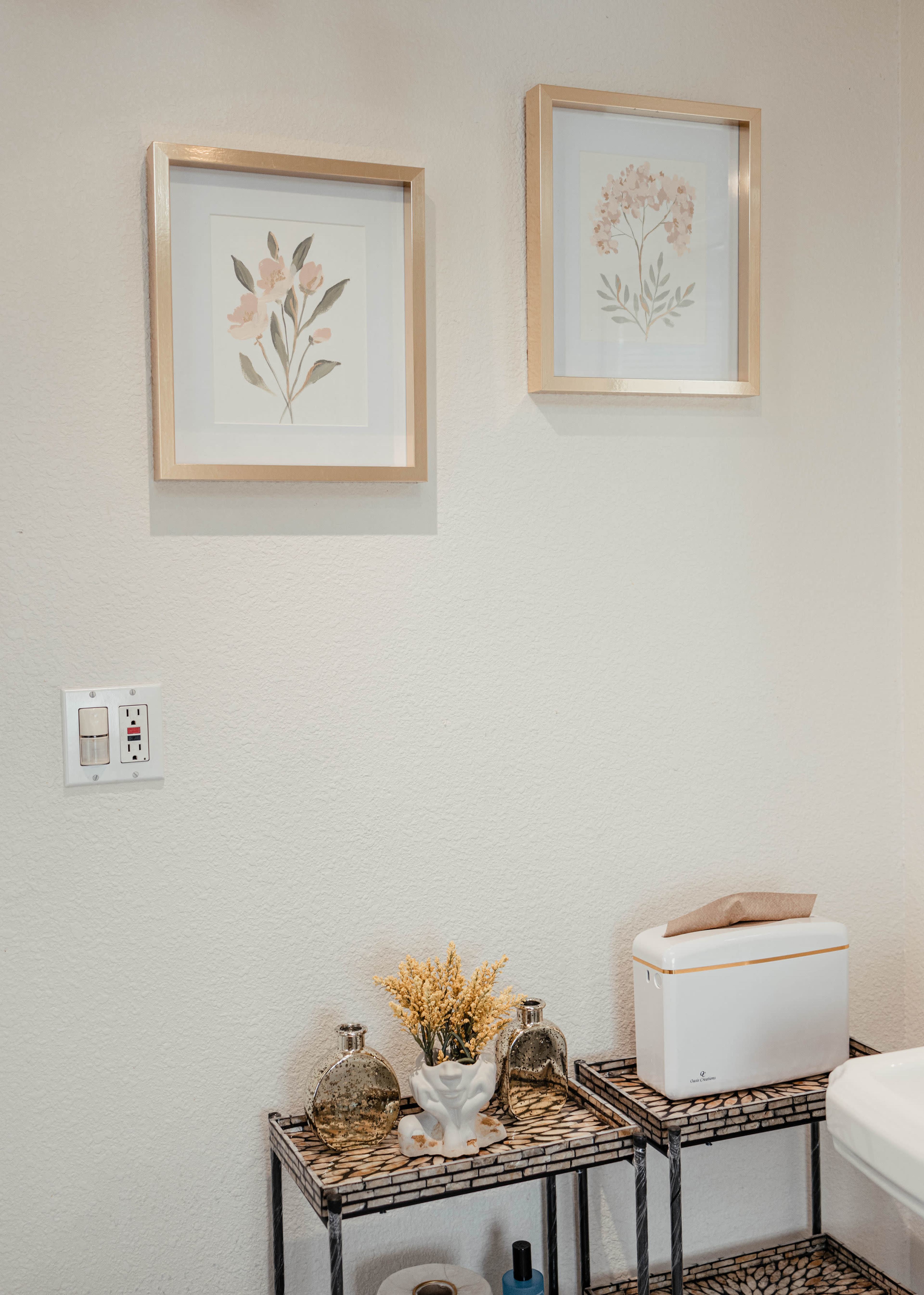 A bathroom scene features two framed botanical prints on the wall above a small shelf with decorative items, including glass bottles and a white container.