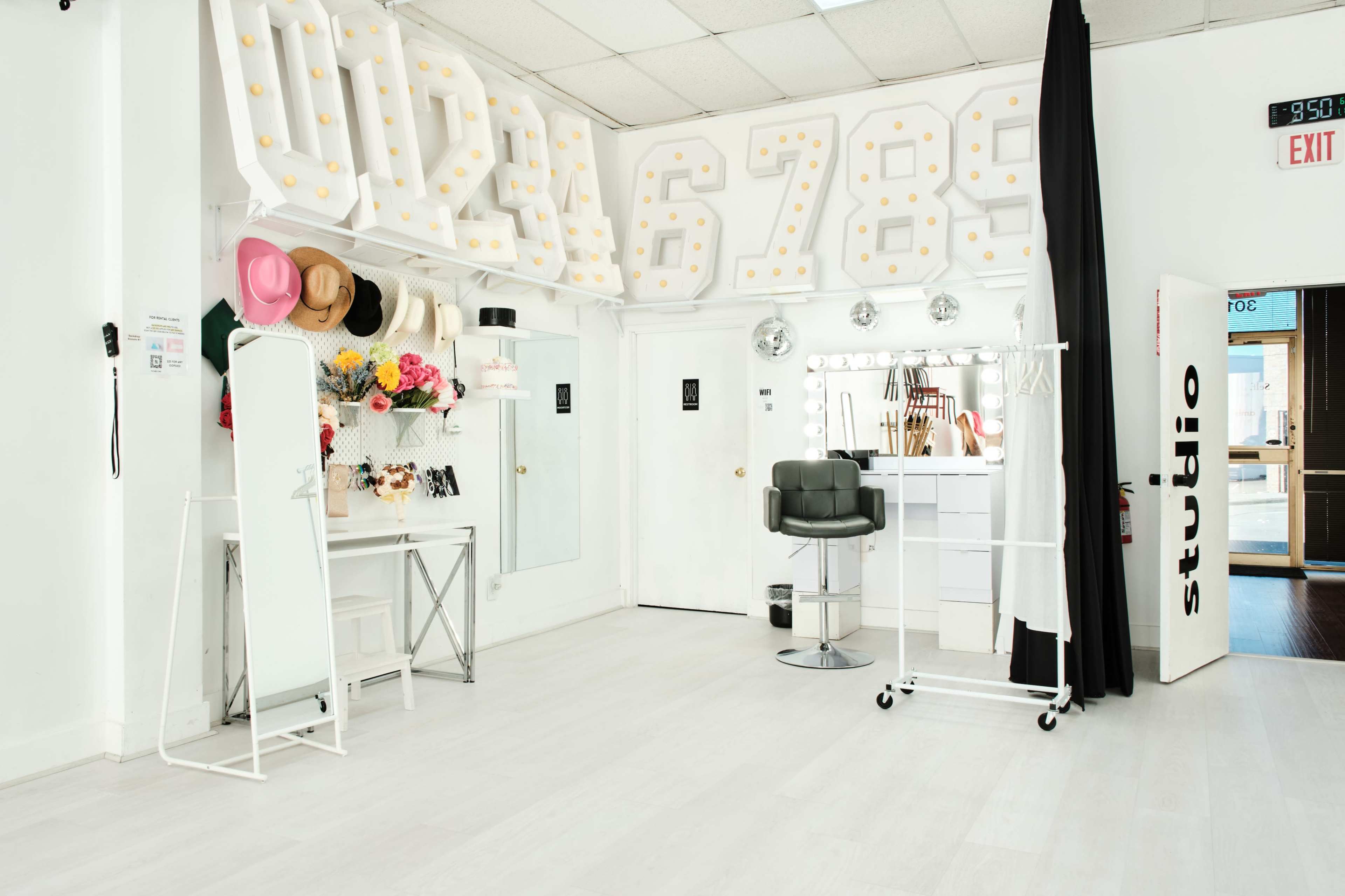A brightly lit salon interior features a mirrored vanity, a styling chair, and decorative hats, with a backdrop of large illuminated numbers on the wall.