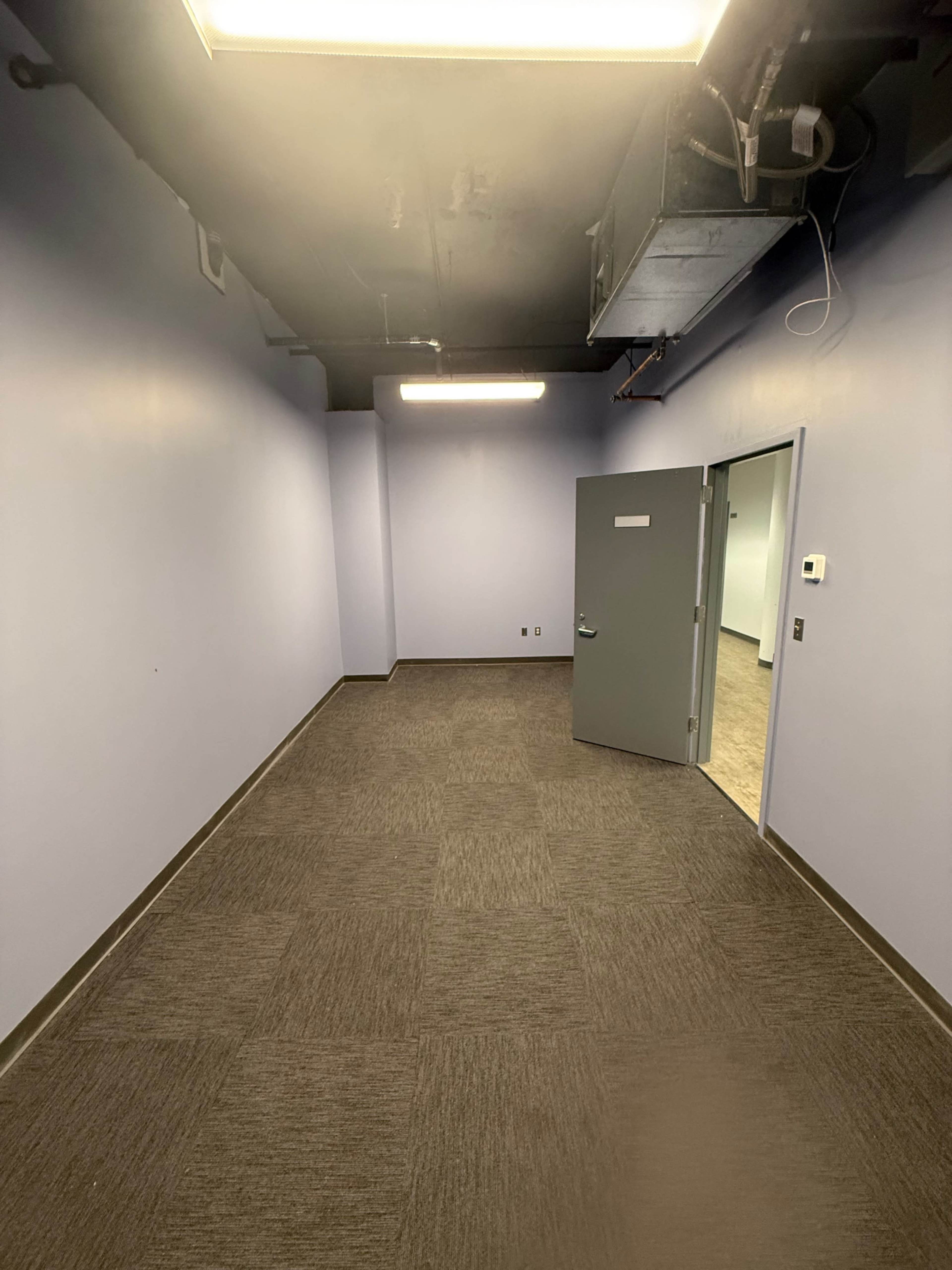 The image shows a vacant room with gray walls, carpeted flooring, and a door leading to another area.