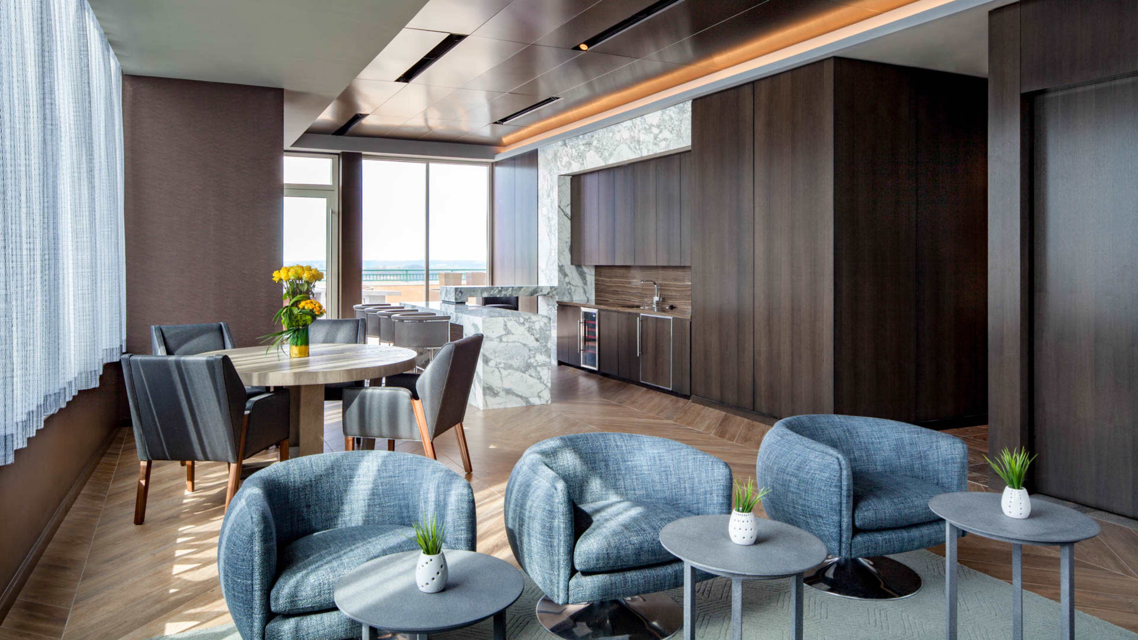 A modern, sunlit interior featuring a dining table with chairs, a kitchenette area, and blue armchairs arranged around small side tables.