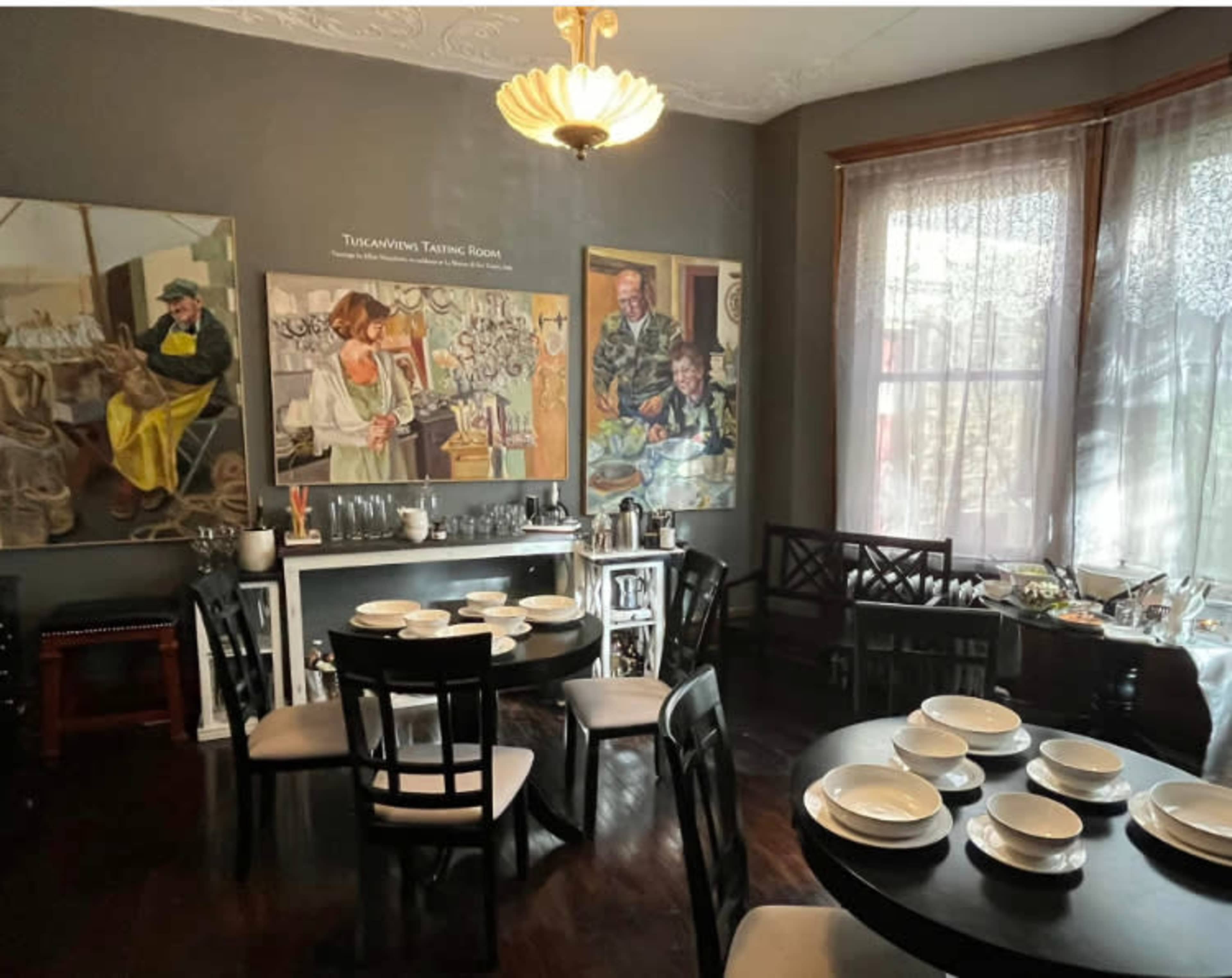 A dining room features black tables set with white plates, a sideboard with serving utensils, and two large paintings on the walls.
