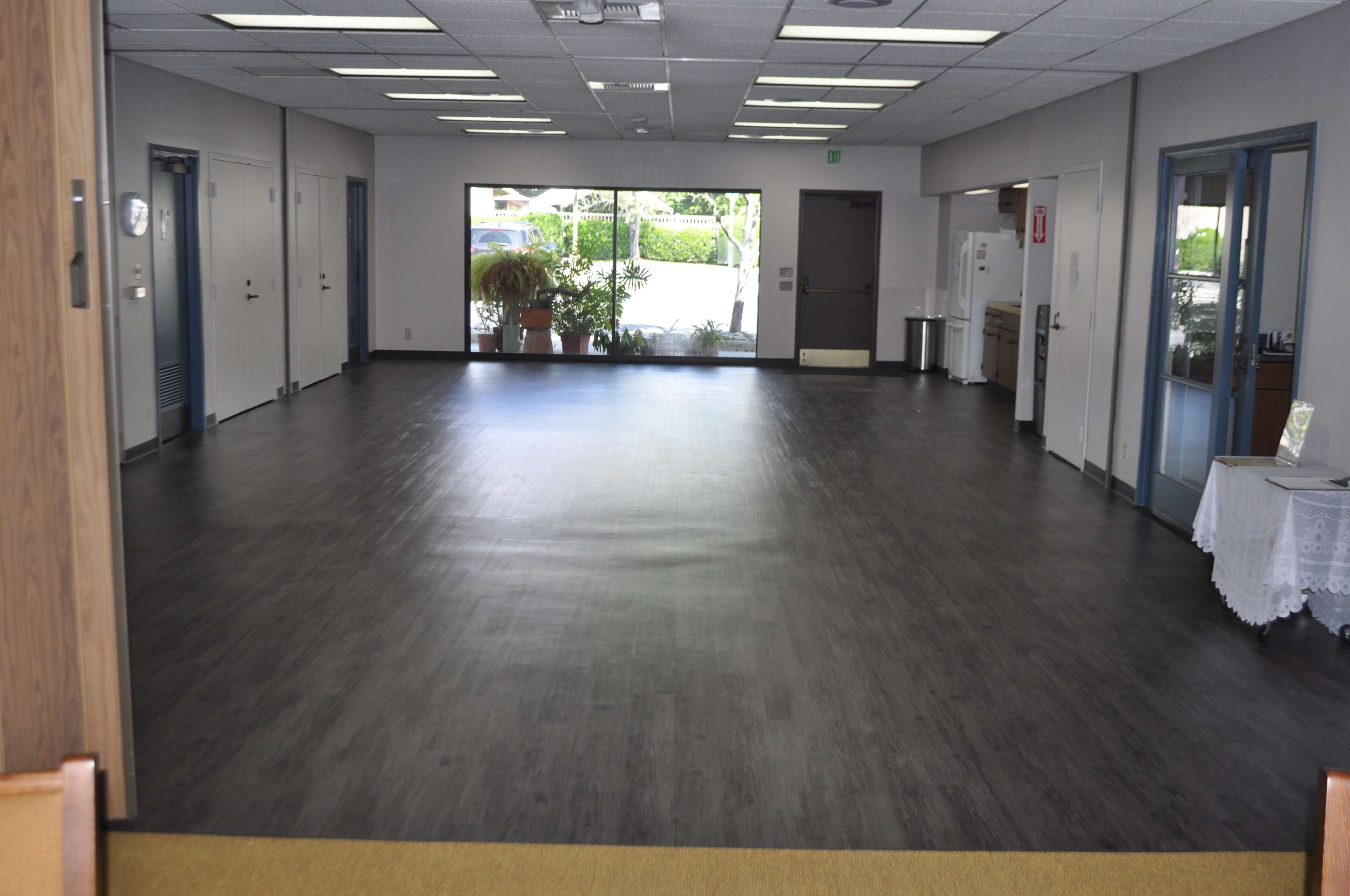 A spacious, empty room with a dark wooden floor, large windows on one side, and a kitchenette visible in the background.