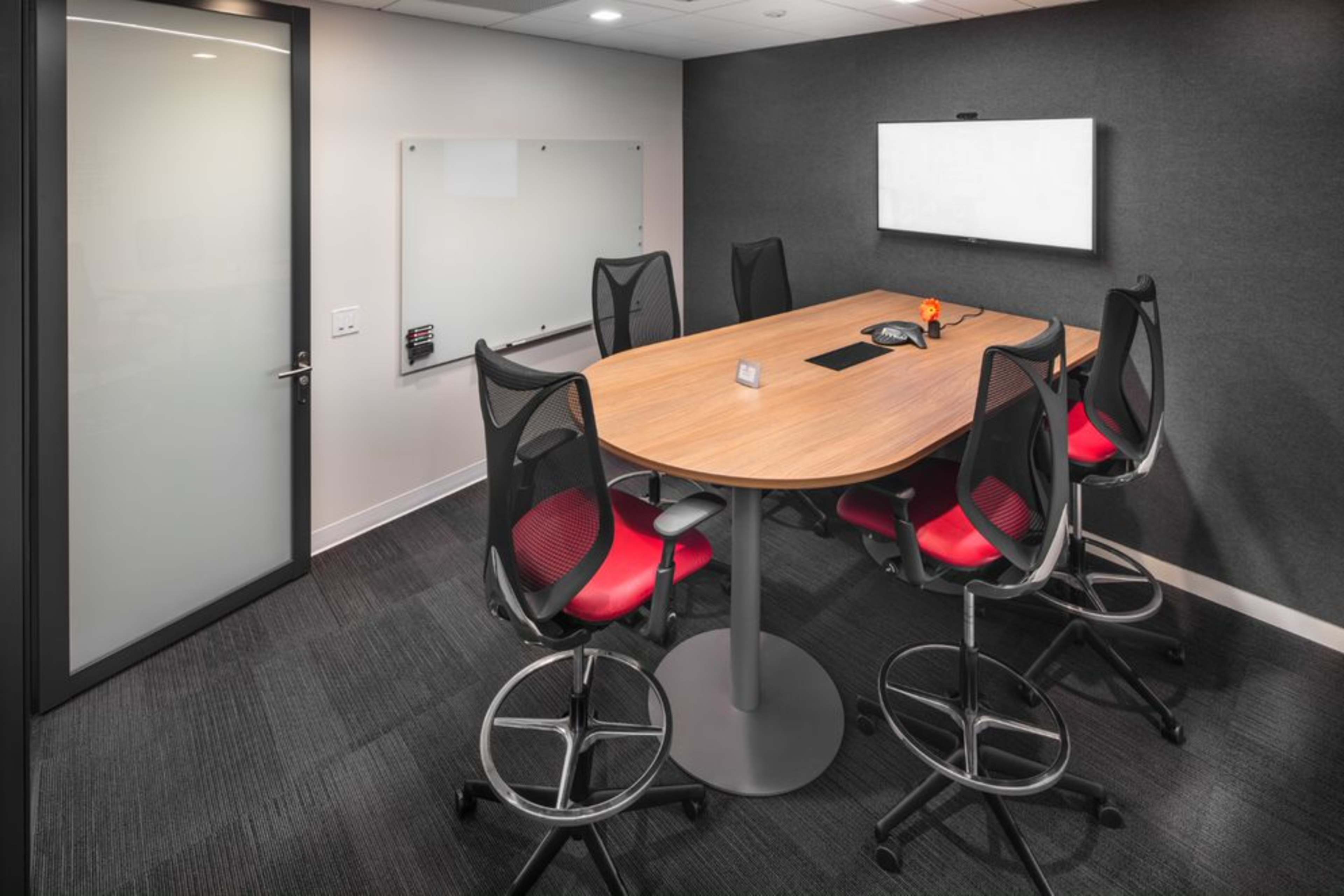 A modern conference room with a round wooden table surrounded by six high-backed chairs and equipped with a wall-mounted display and a whiteboard.