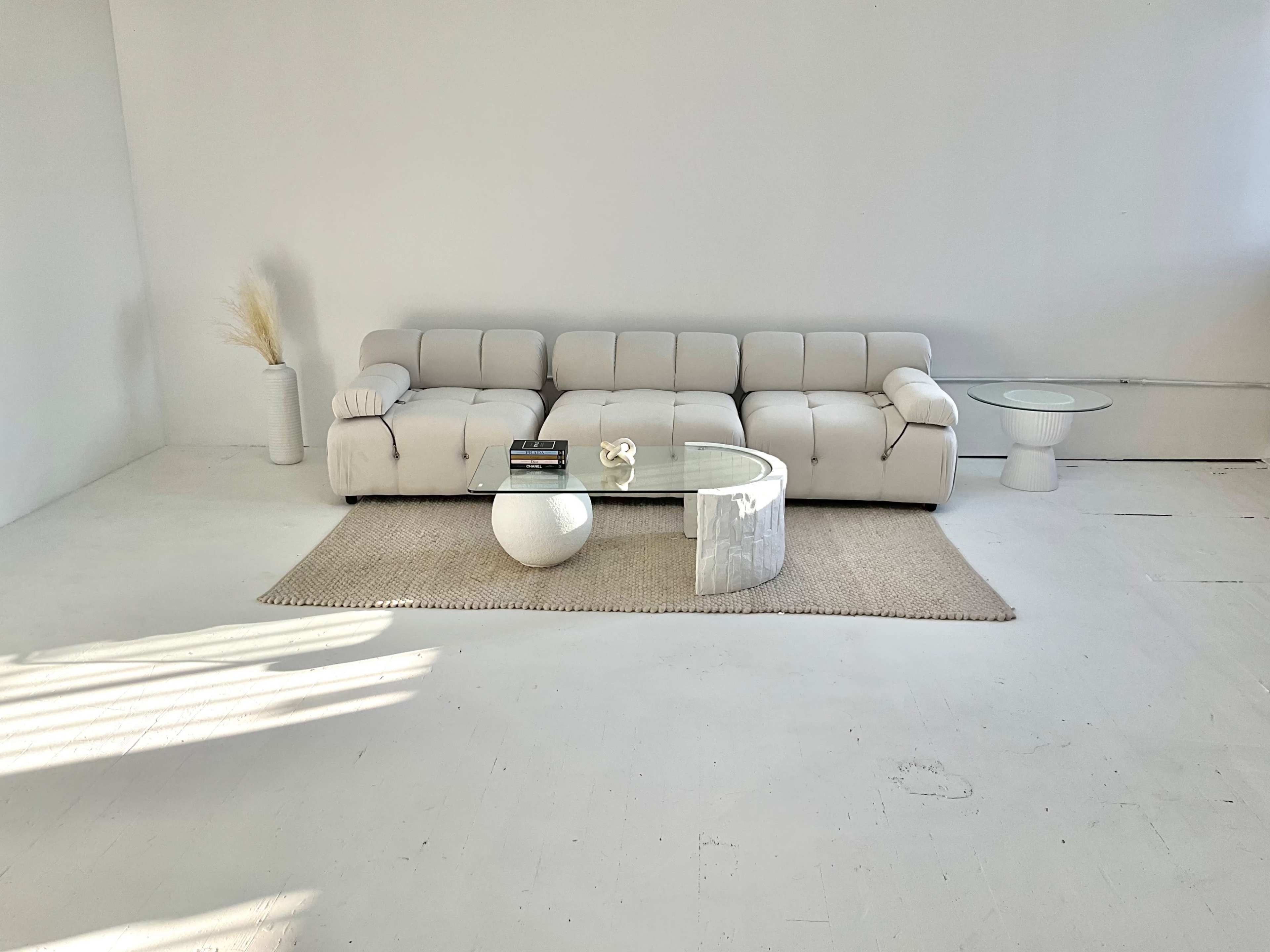 A light-colored sectional sofa is placed on a woven rug in a minimalist room, accompanied by a glass coffee table and a small white side table.