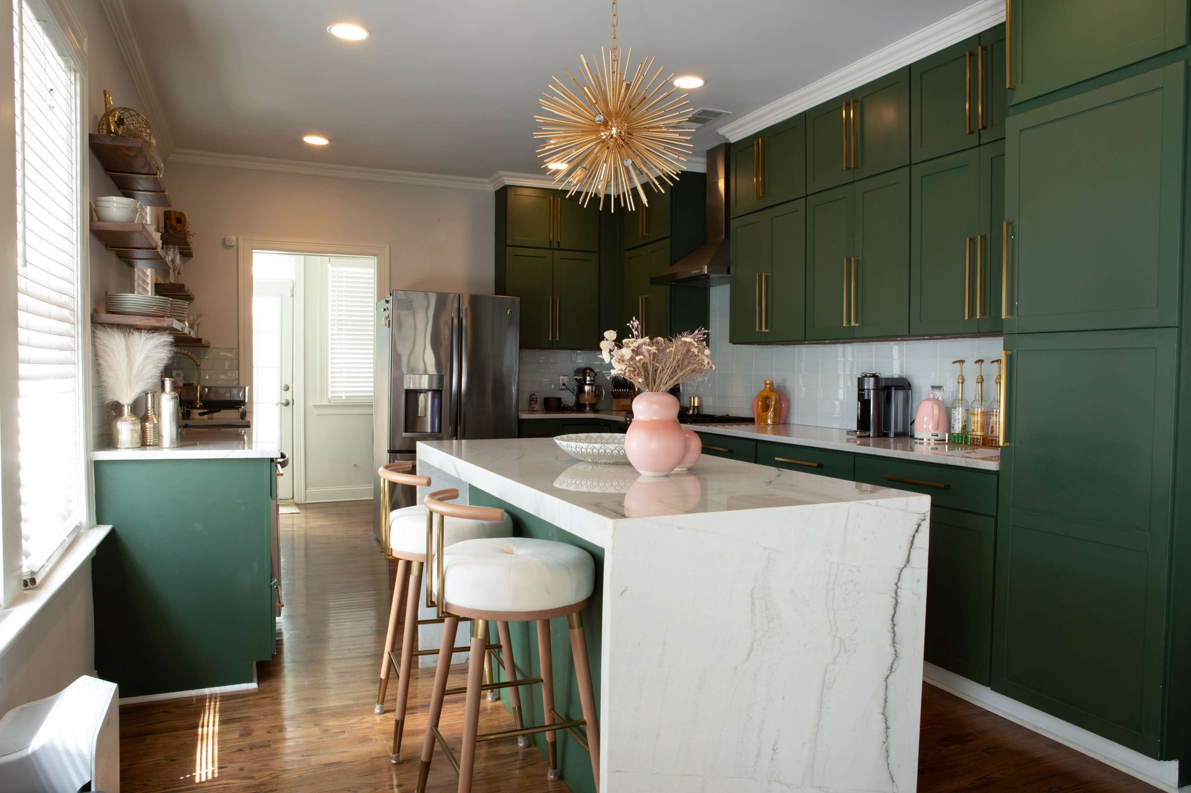 The image shows a modern kitchen with dark green cabinets, a marbled white countertop, a gold starburst pendant light, and wooden flooring.