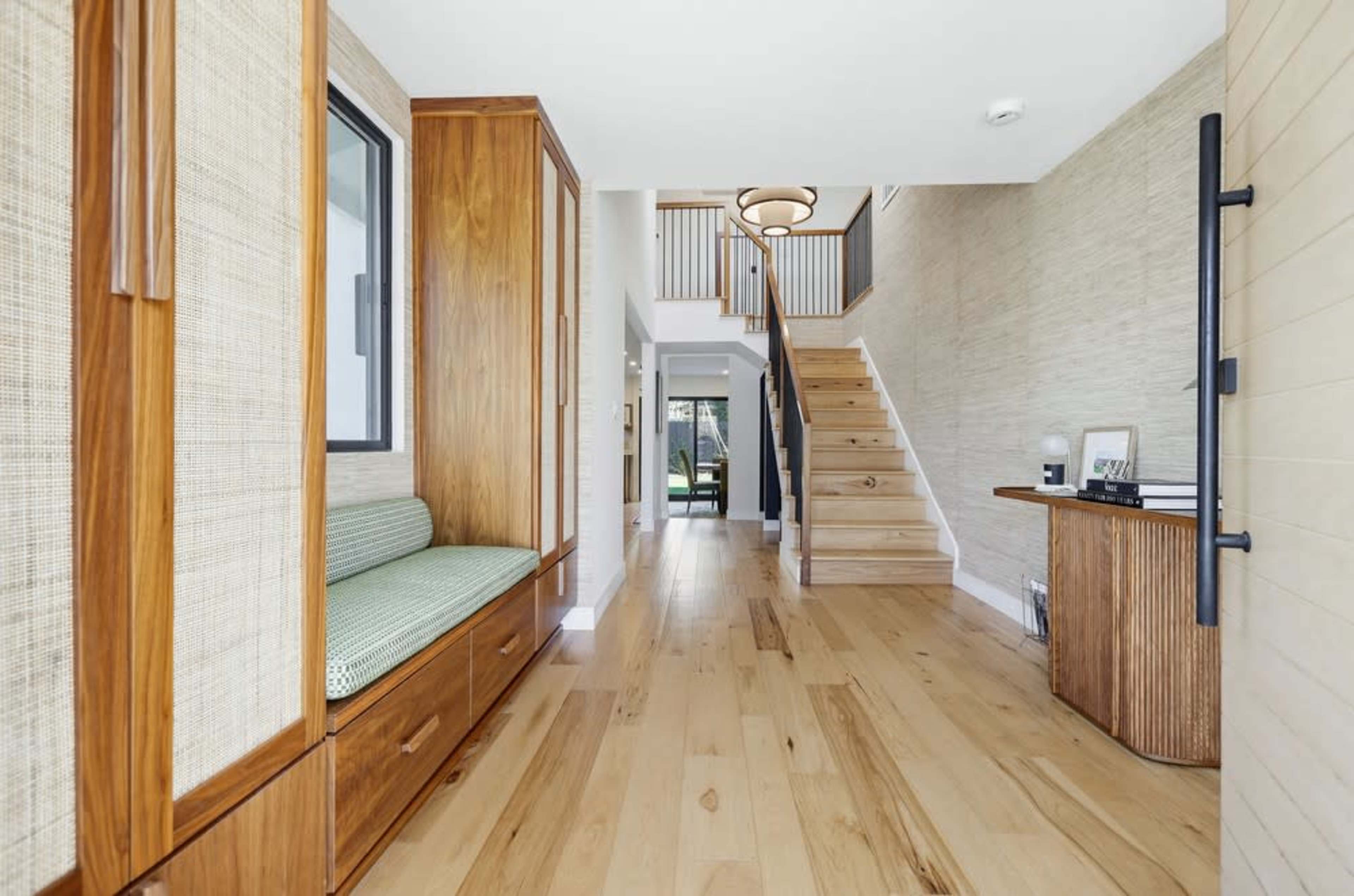 The scene features a modern foyer with wooden flooring, a staircase, a built-in bench, and natural lighting.
