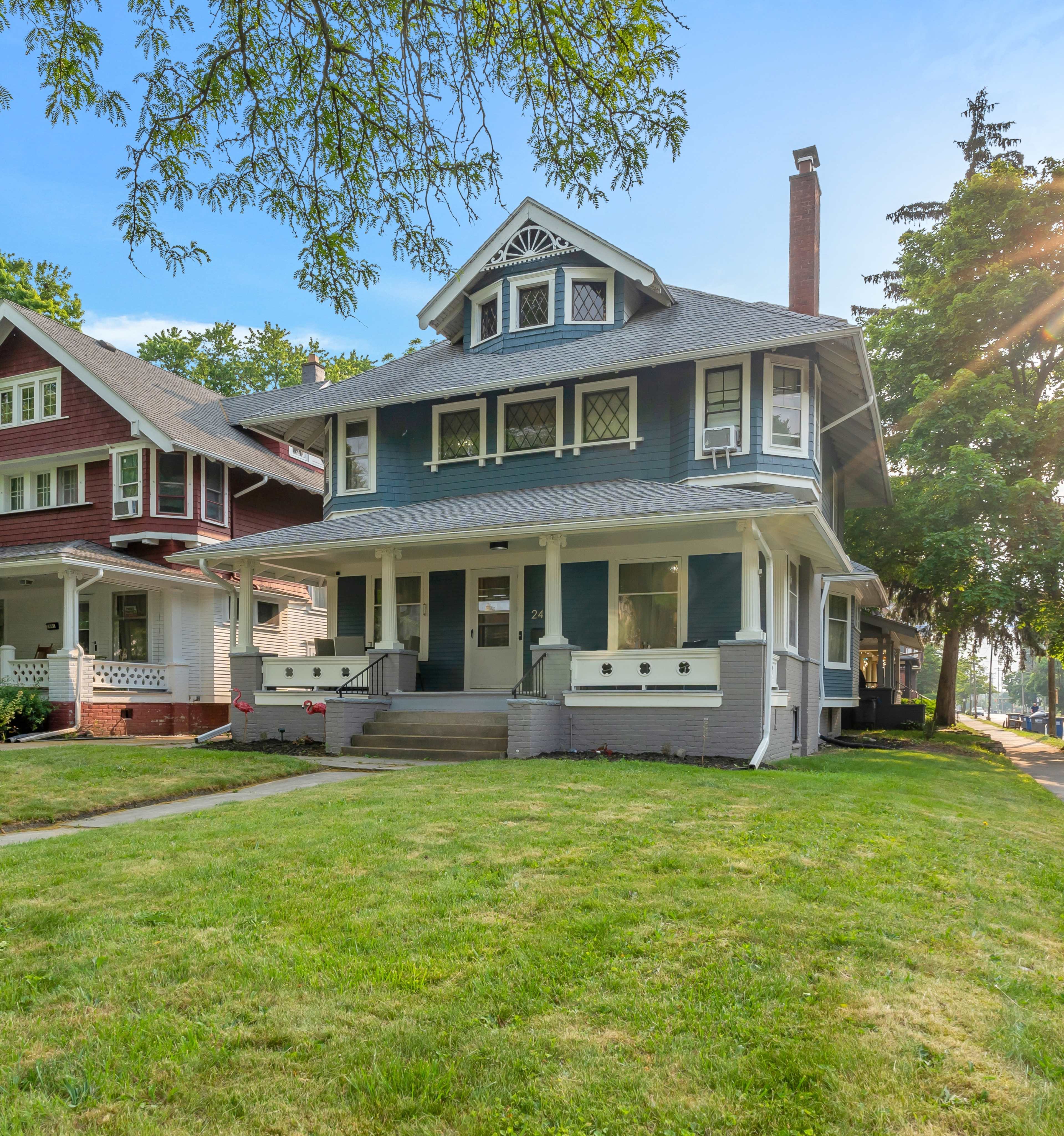 A two-story, dark blue house with a porch and decorative gables, situated on a grassy lawn alongside other houses.